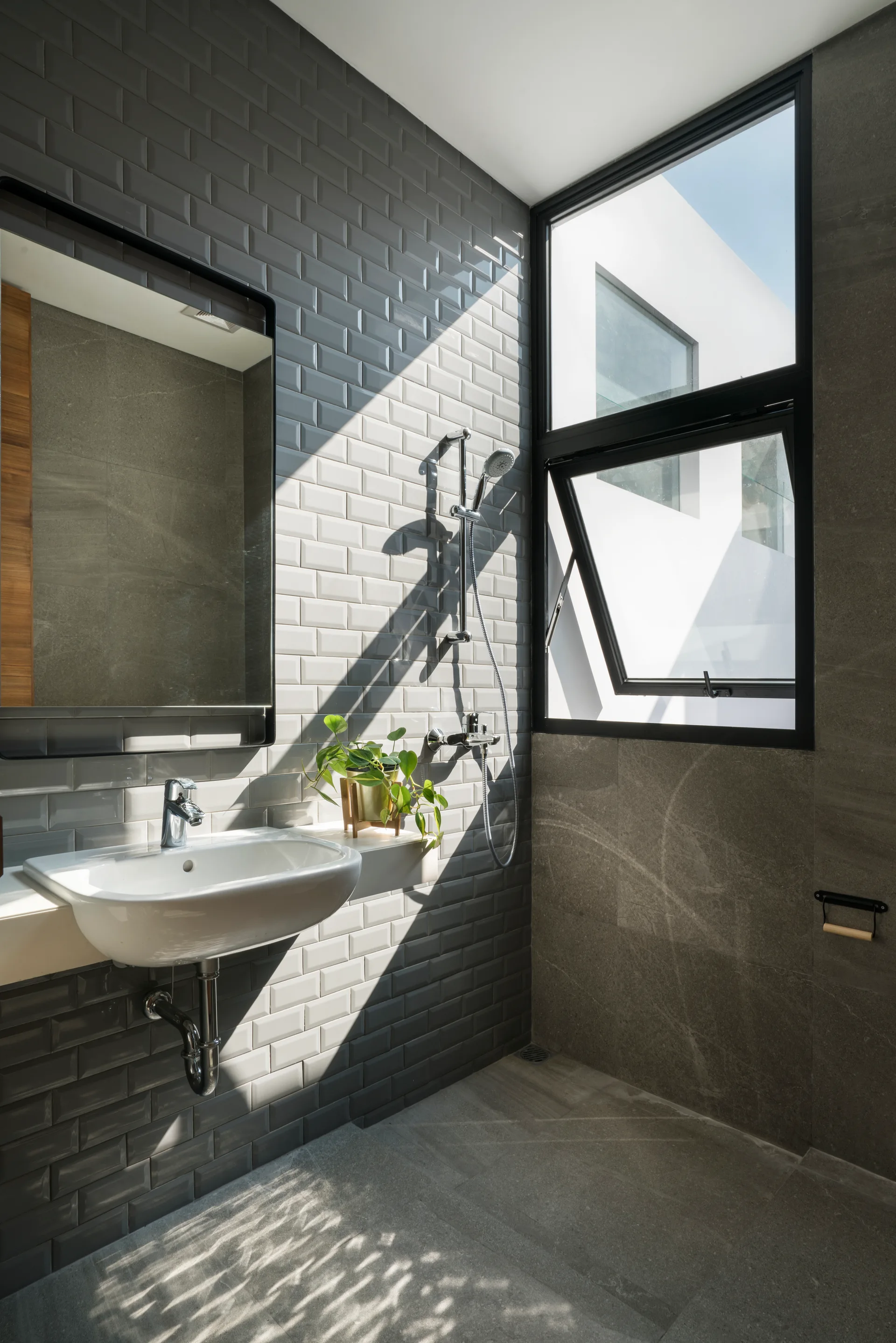 A modern bathroom with glossy grey subway tile walls, a wall-mounted white ceramic sink, and a large black-framed window letting in sunlight.