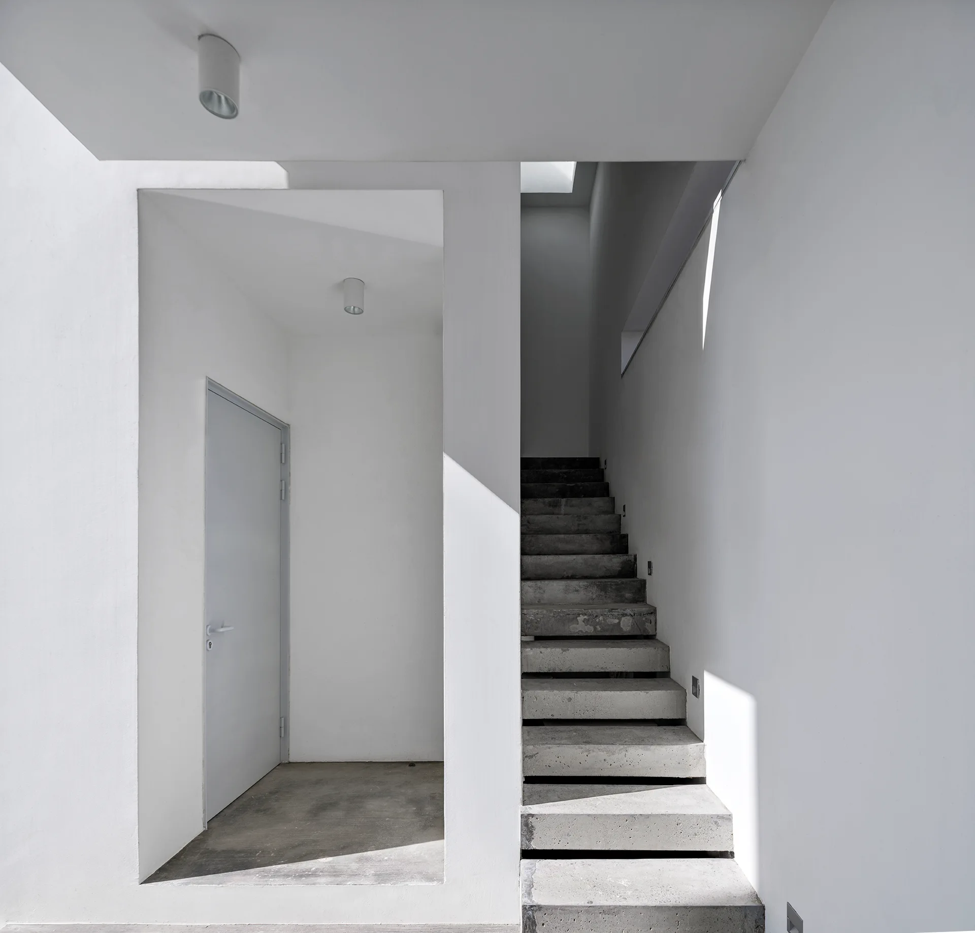 A minimalist concrete staircase rises next to a white plastered entryway with a simple door and geometric shadows.