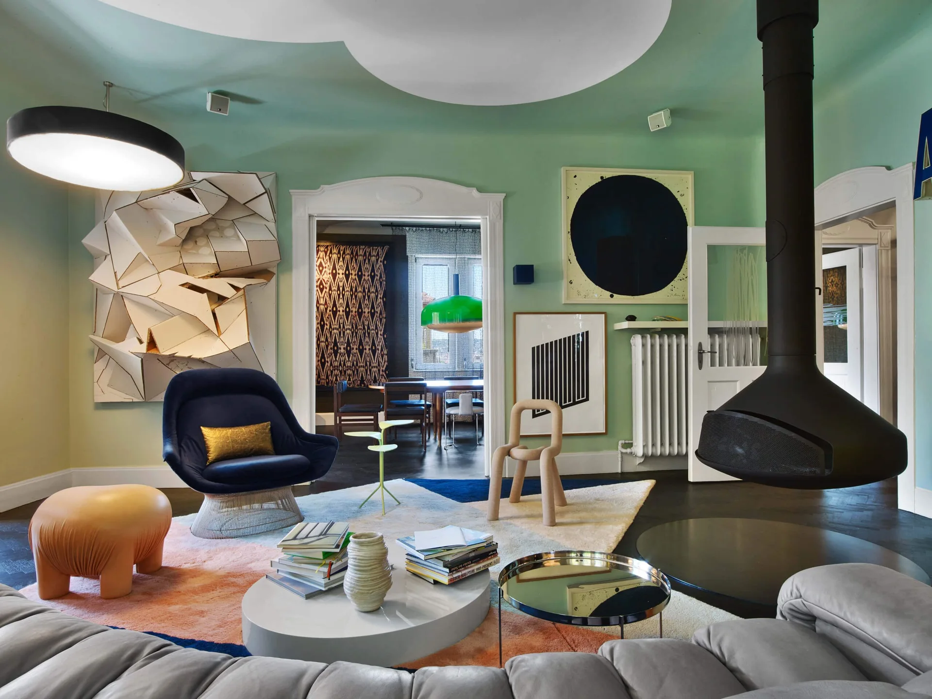An eclectic living room with mint green walls, a black suspended fireplace, colorful geometric rug, and sculptural lounge chairs.