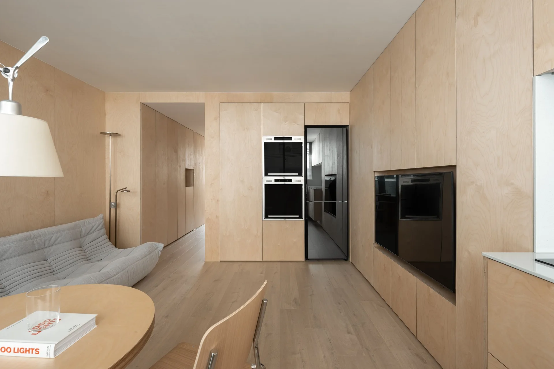 A minimalist room features light plywood wall paneling, a grey low-profile sofa, a round wooden table, and integrated black kitchen appliances.