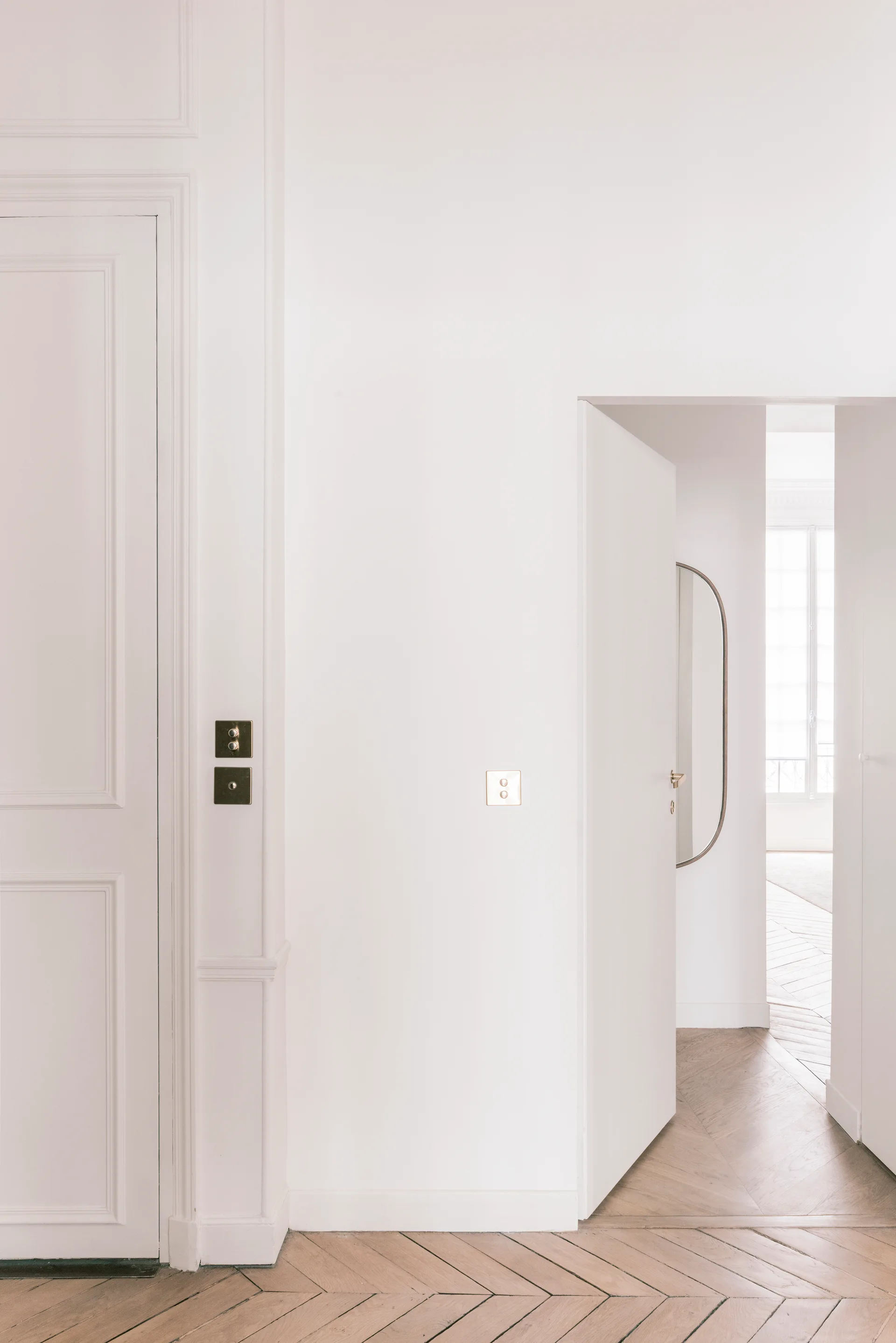 A minimalist white hallway featuring light wood herringbone flooring and traditional wall moldings.