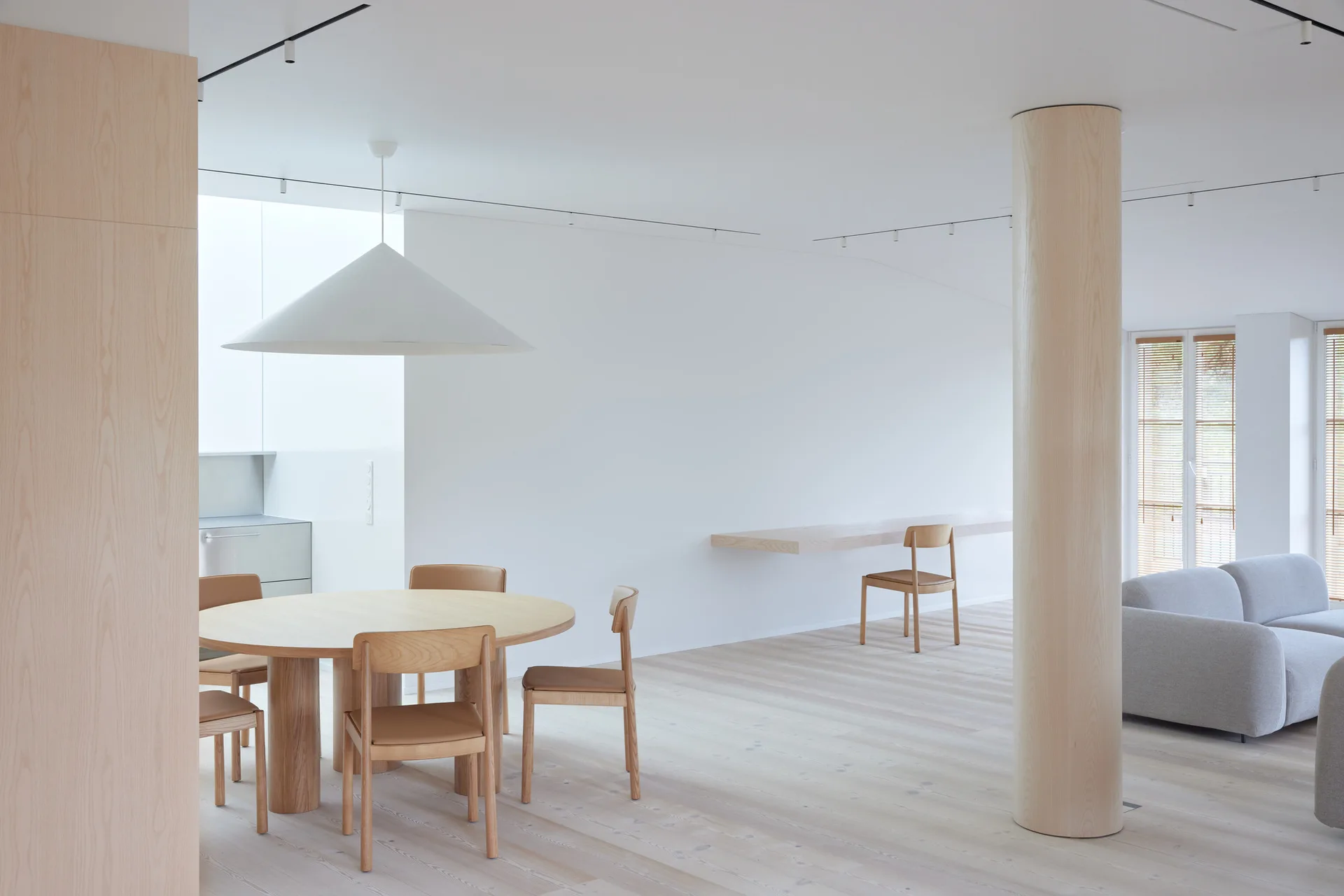 A minimalist open-plan room with a round wooden dining table, a matching cylindrical pillar, and a floating desk along a plain white wall.