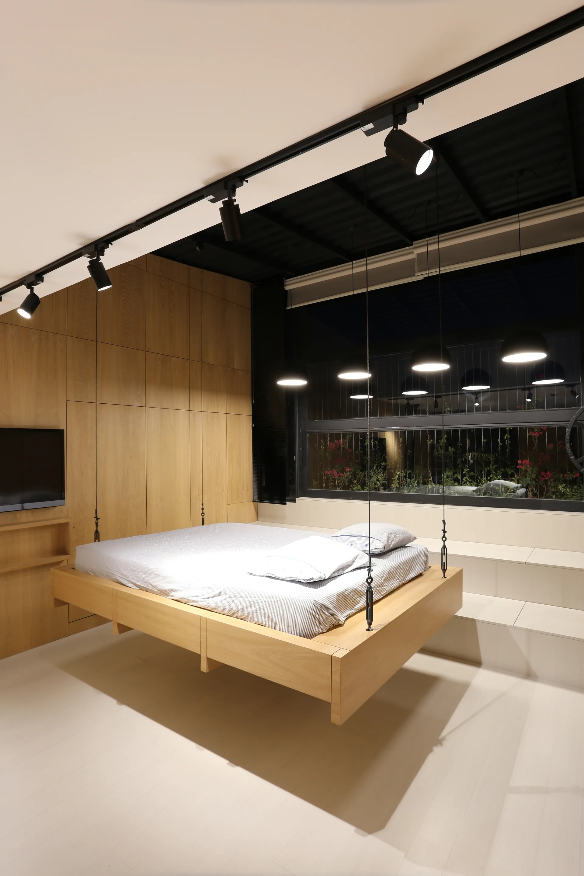 A minimalist bedroom features a light wood bed suspended from the ceiling by thin chains, set against matching built-in oak storage cabinets.