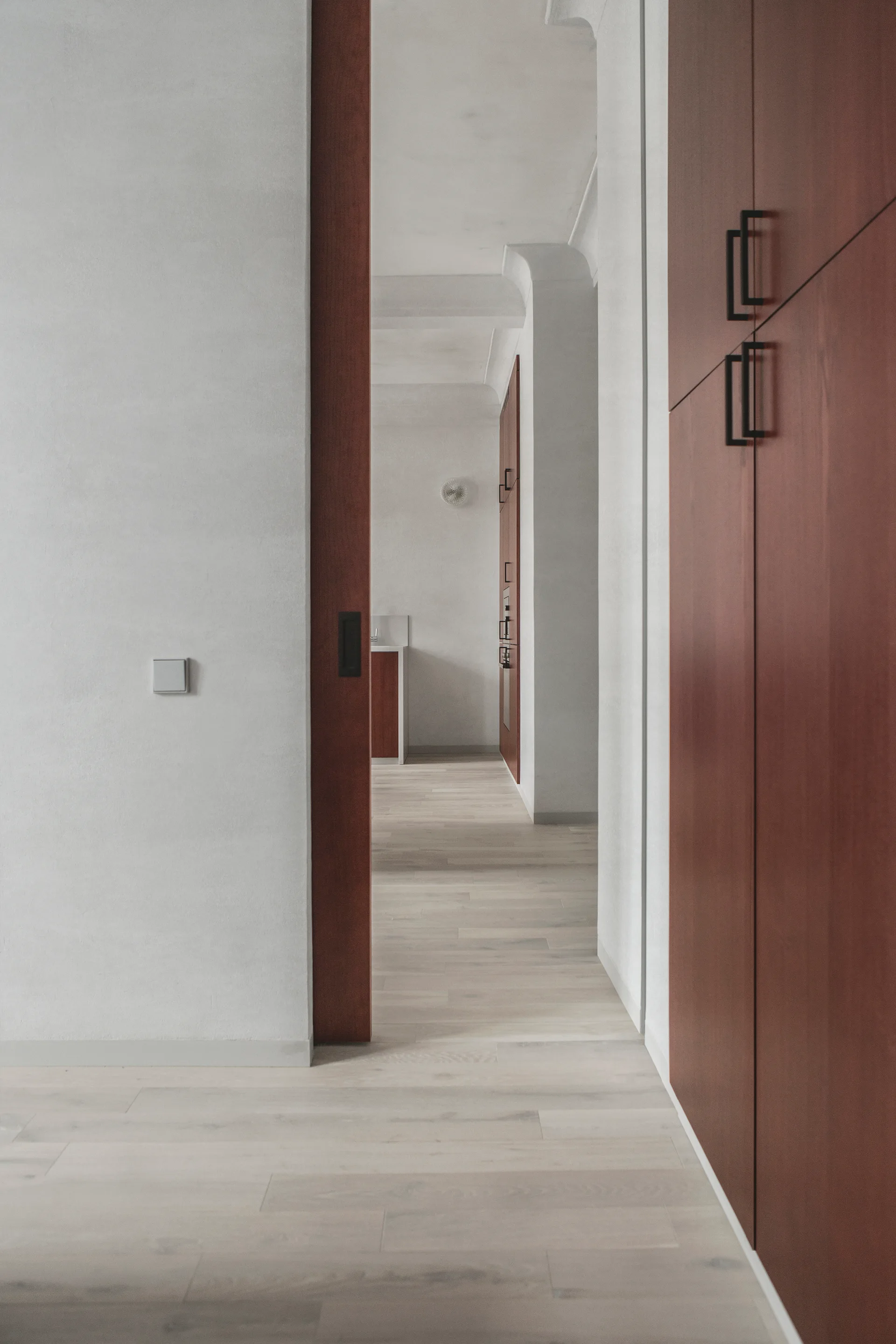 A minimalist hallway features rich reddish-brown wood cabinetry and a matching doorway leading into a bright kitchen with light oak floors.