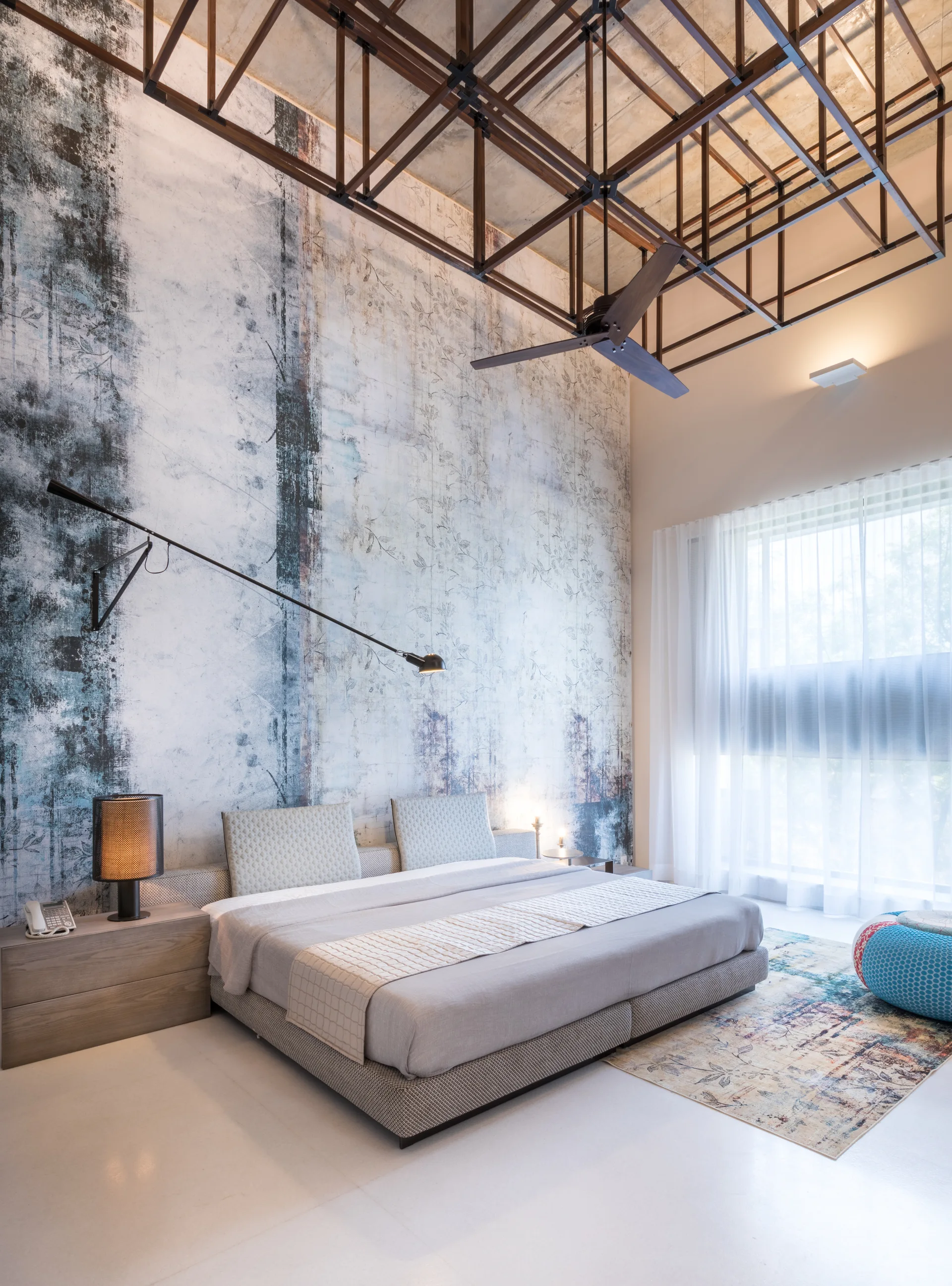 A contemporary bedroom features a low platform bed set against a tall abstract mural wall beneath a dark metal ceiling grid structure.