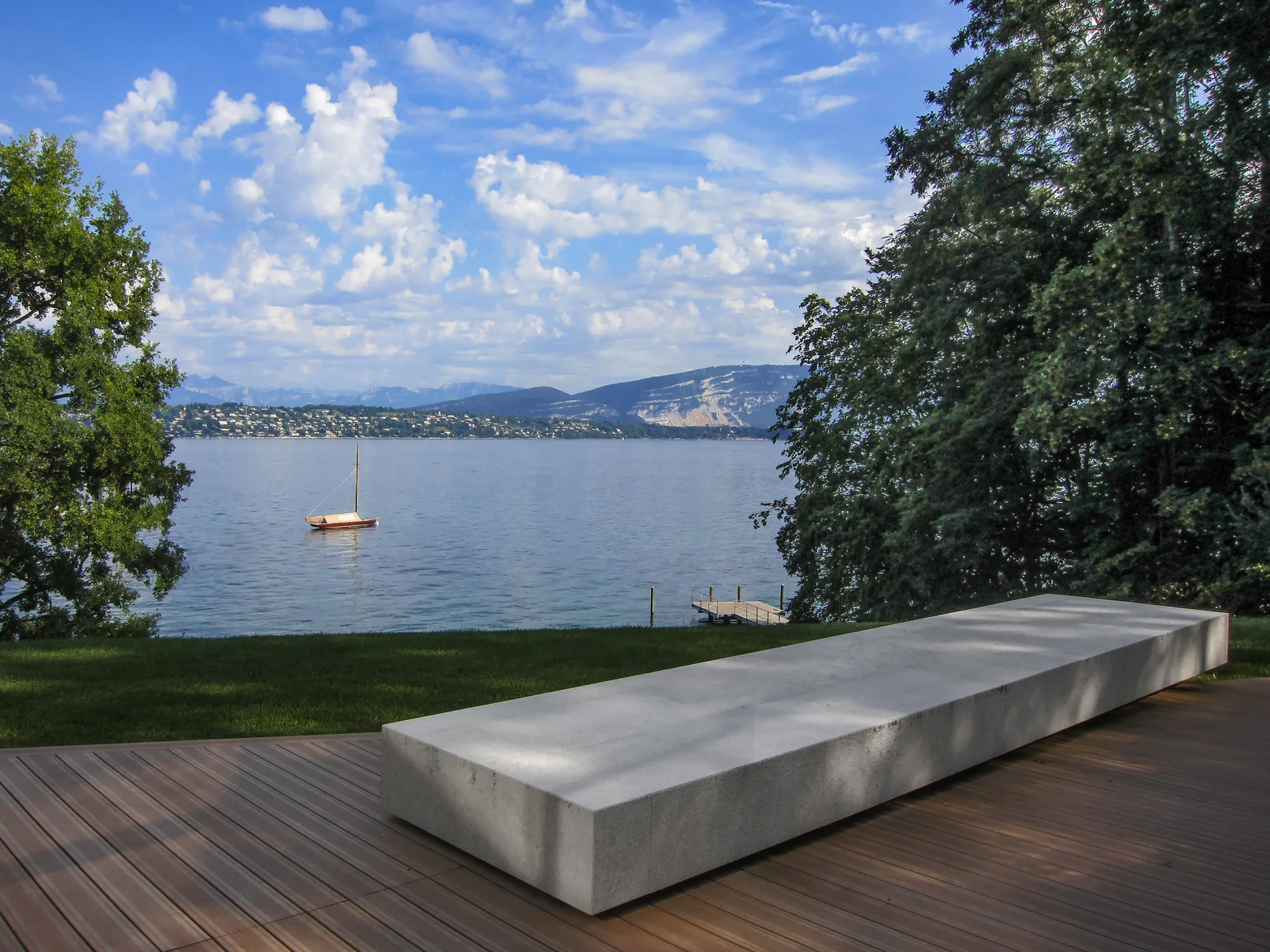 A long rectangular concrete bench sits on a wooden deck overlooking a wide lake with mountains in the background.