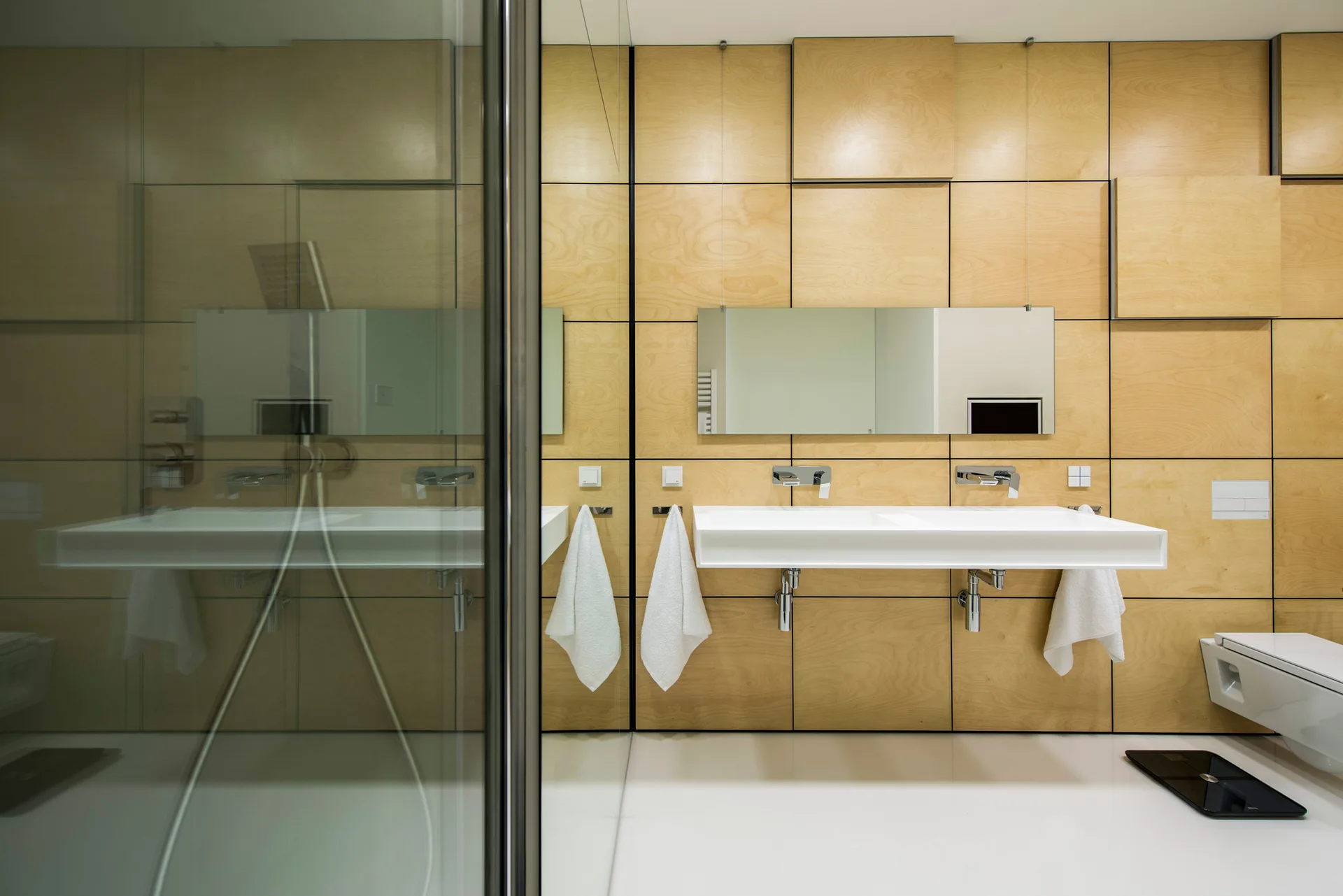 A modern minimalist bathroom featuring a gridded light wood paneled wall, a white wall-mounted double sink, and a glass shower enclosure.