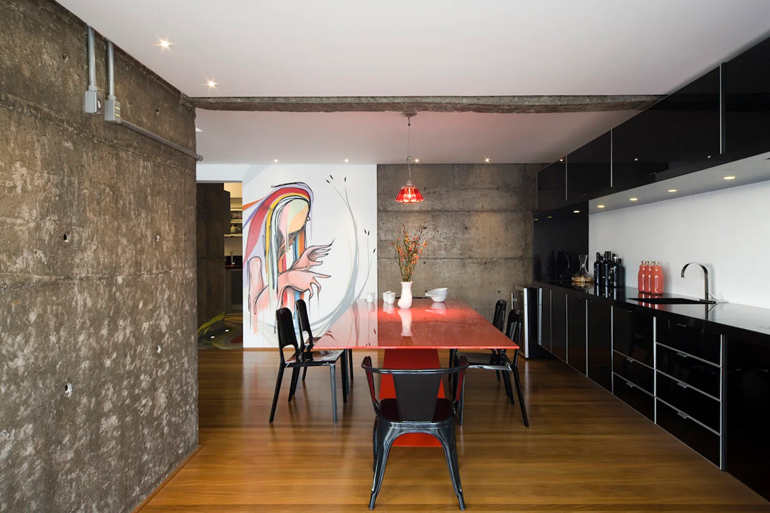 A modern dining room features a glossy red table and black chairs set against textured raw concrete walls.