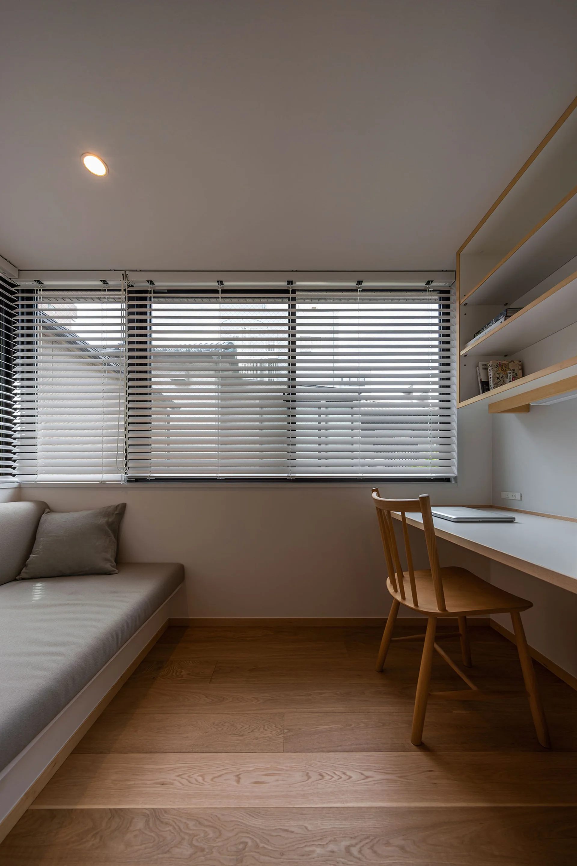 A minimalist home office features a built-in white desk with a wooden chair, a low daybed with a grey cushion, and a window with horizontal blinds.