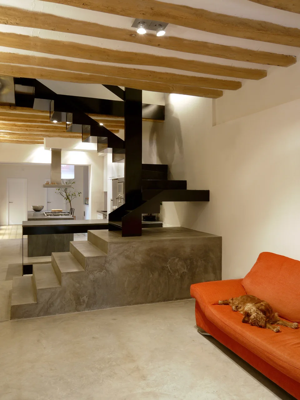 A modern living room features a black metal staircase on a concrete base, exposed wooden ceiling beams, concrete floors, and an orange sofa.