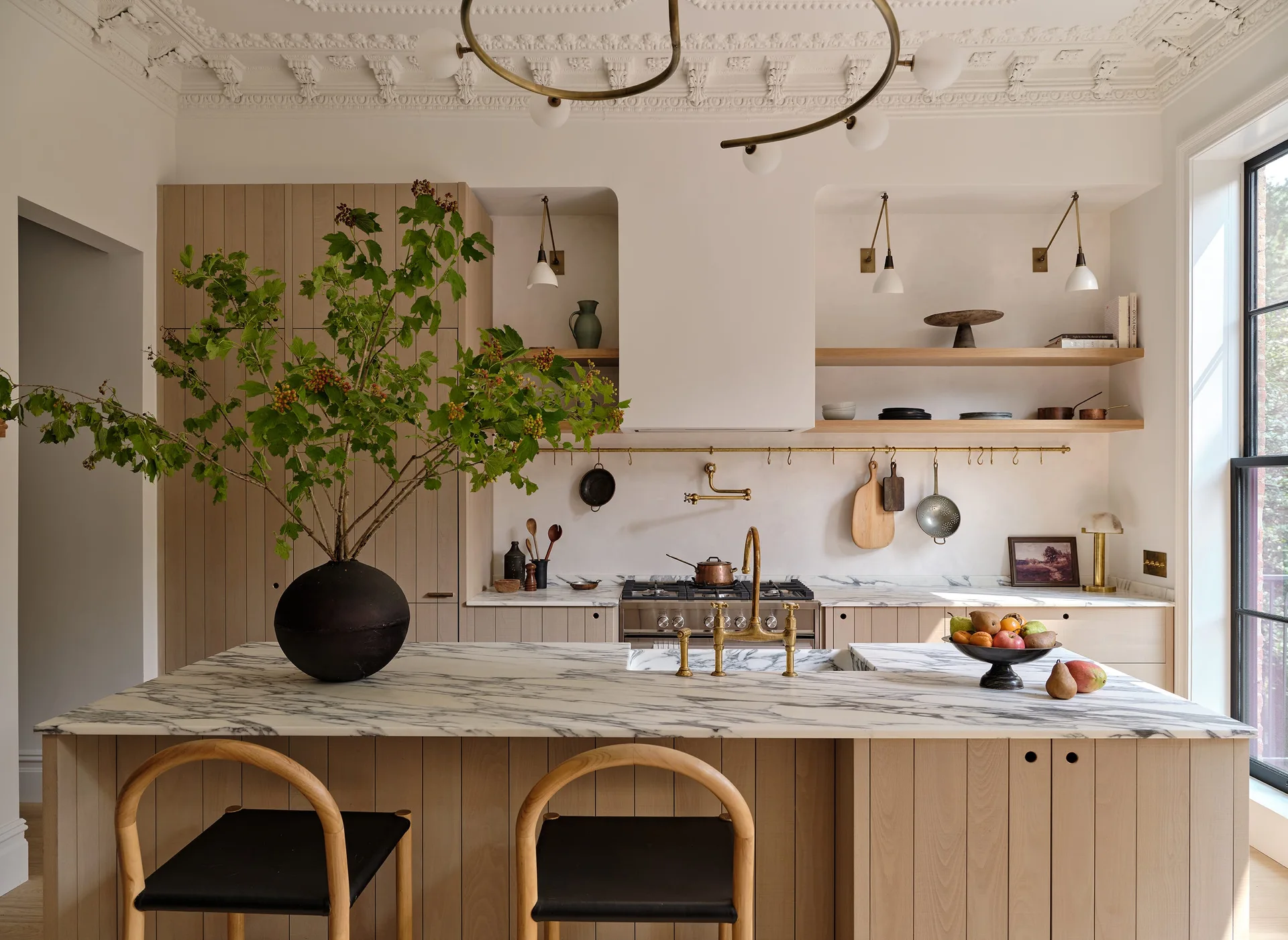 A kitchen features a marble-topped island with two wooden bar stools, vertical slatted wood cabinetry, and intricate white ceiling molding.