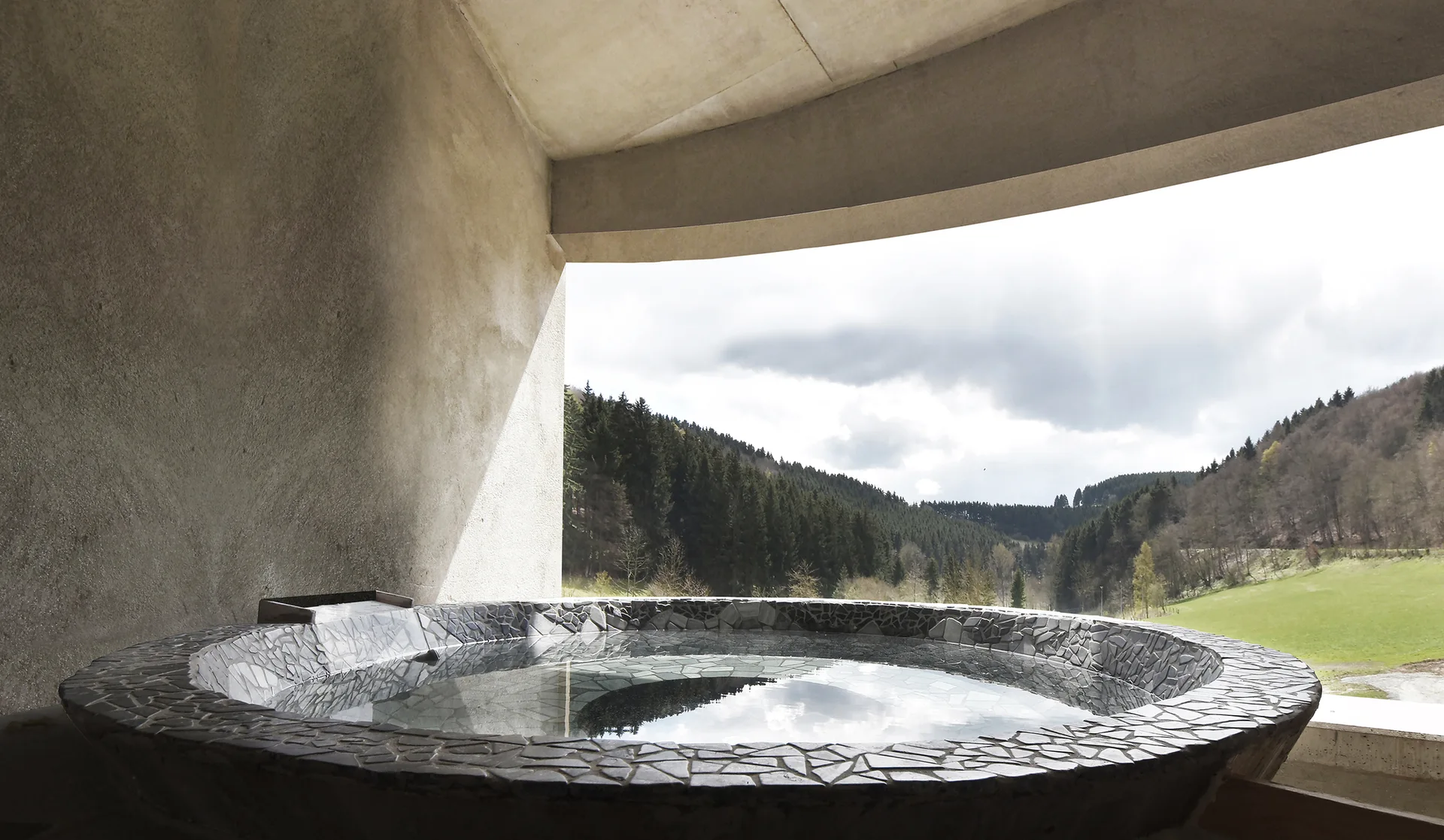A circular soaking tub with a grey stone mosaic rim sits under a curved concrete shelter overlooking a forested valley.