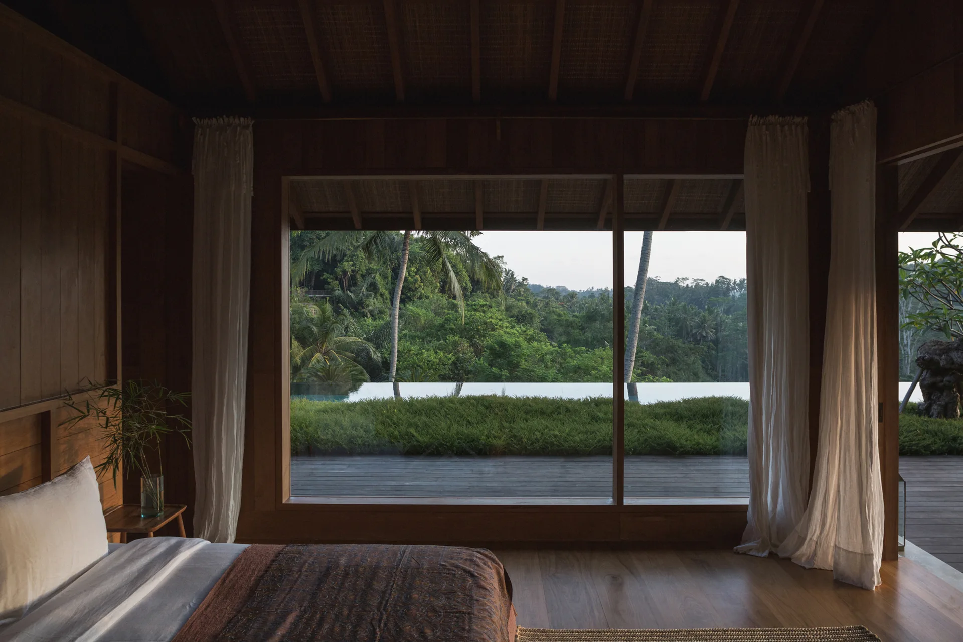 A wooden bedroom features a low bed, sheer white curtains, and a large window framing a lush green tropical landscape.