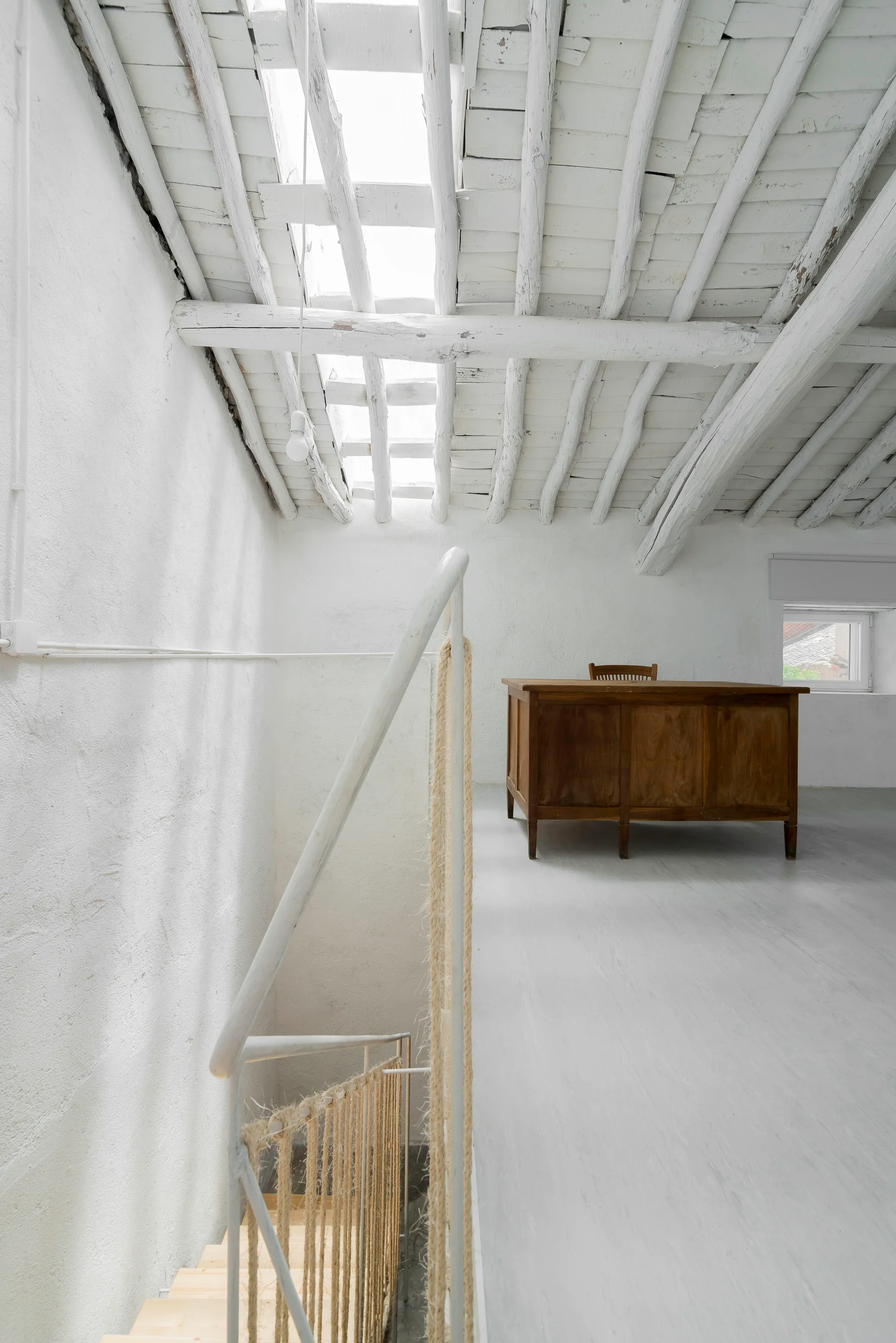 A white attic loft landing features a rustic wooden desk, exposed painted ceiling beams, a skylight, and a staircase railing with rope details.
