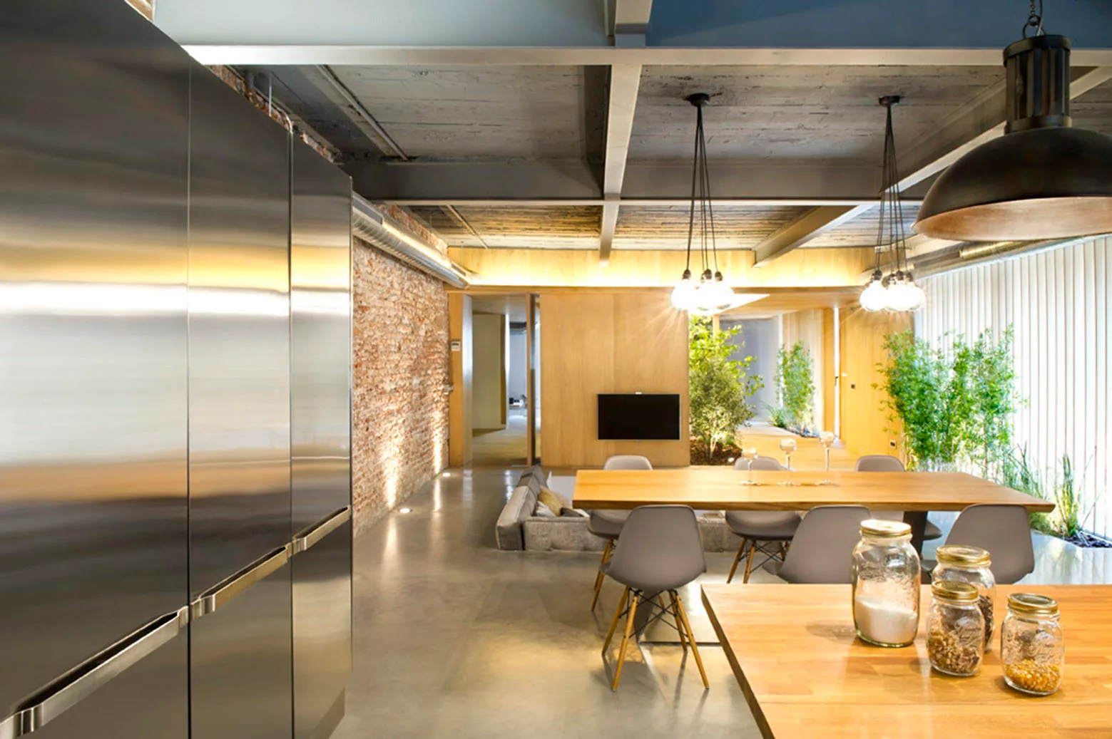 An industrial loft kitchen and dining area featuring a stainless steel refrigerator, a wooden dining table, an exposed brick wall, and a concrete ceiling.