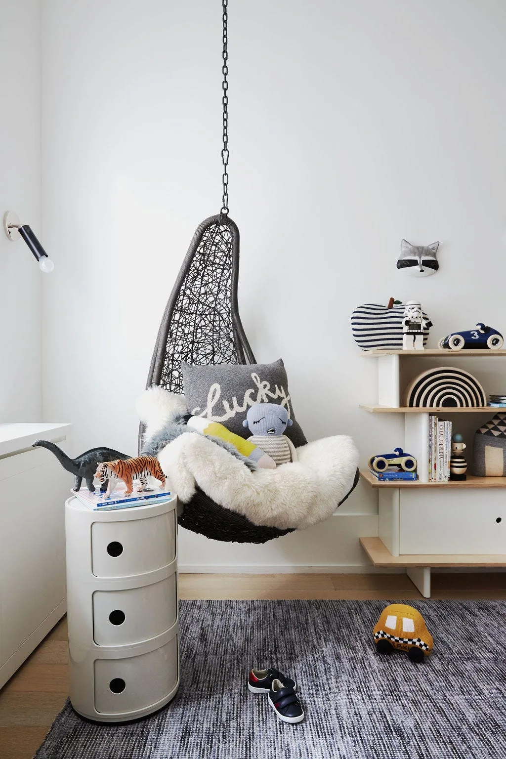 A children's room features a black woven hanging chair with a plush throw next to a white cylindrical storage unit and a wooden toy shelf.