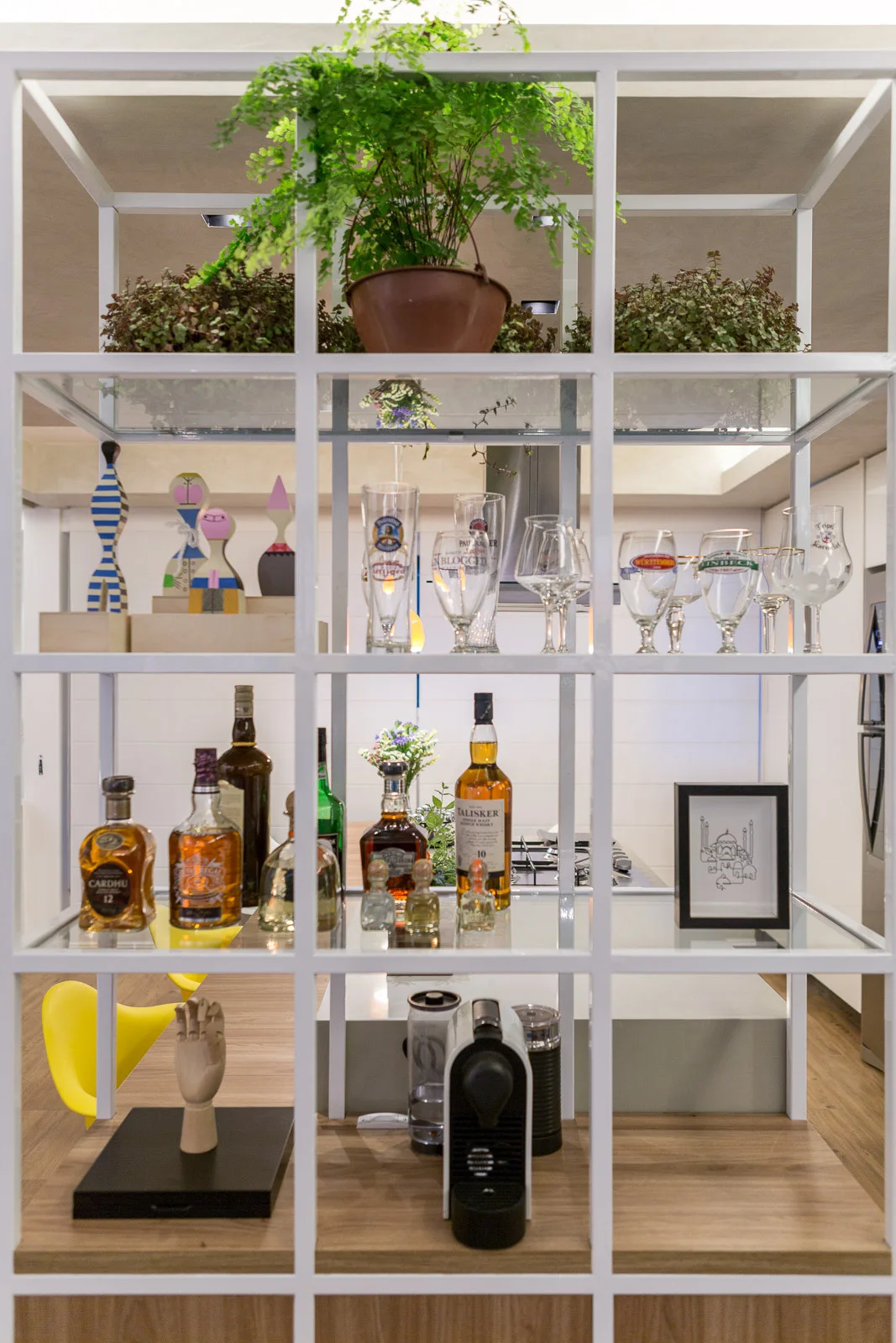 A white metal open shelving unit displays liquor bottles and glassware, dividing a kitchen area with yellow bar stools in the background.