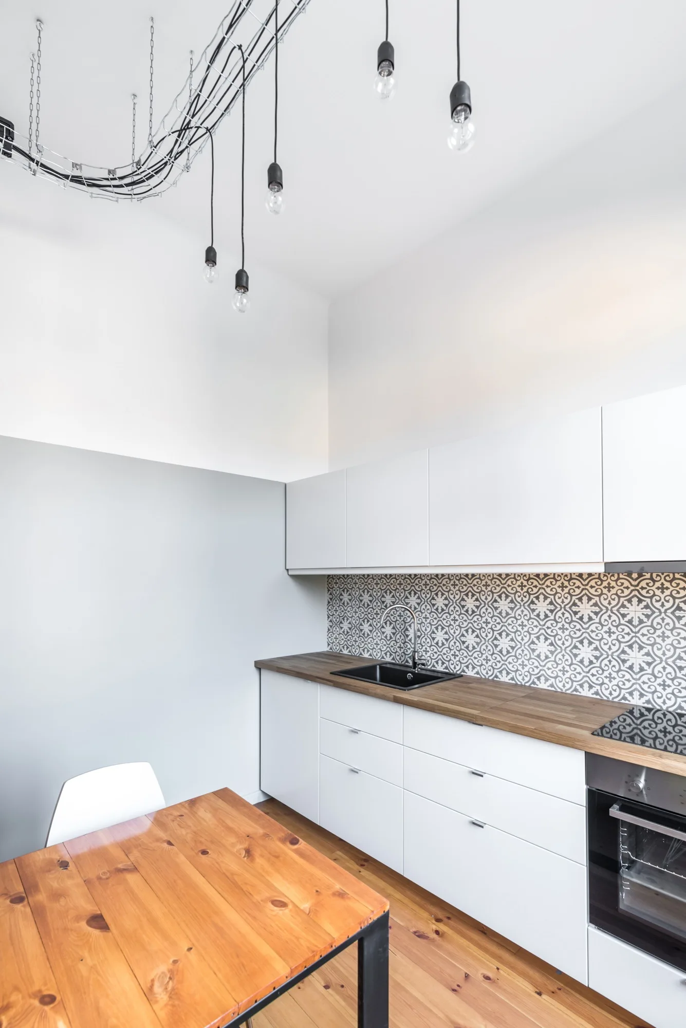 A modern kitchen features white flat-front cabinets, a geometric patterned tile backsplash, a wood countertop, and industrial hanging bulb lights.