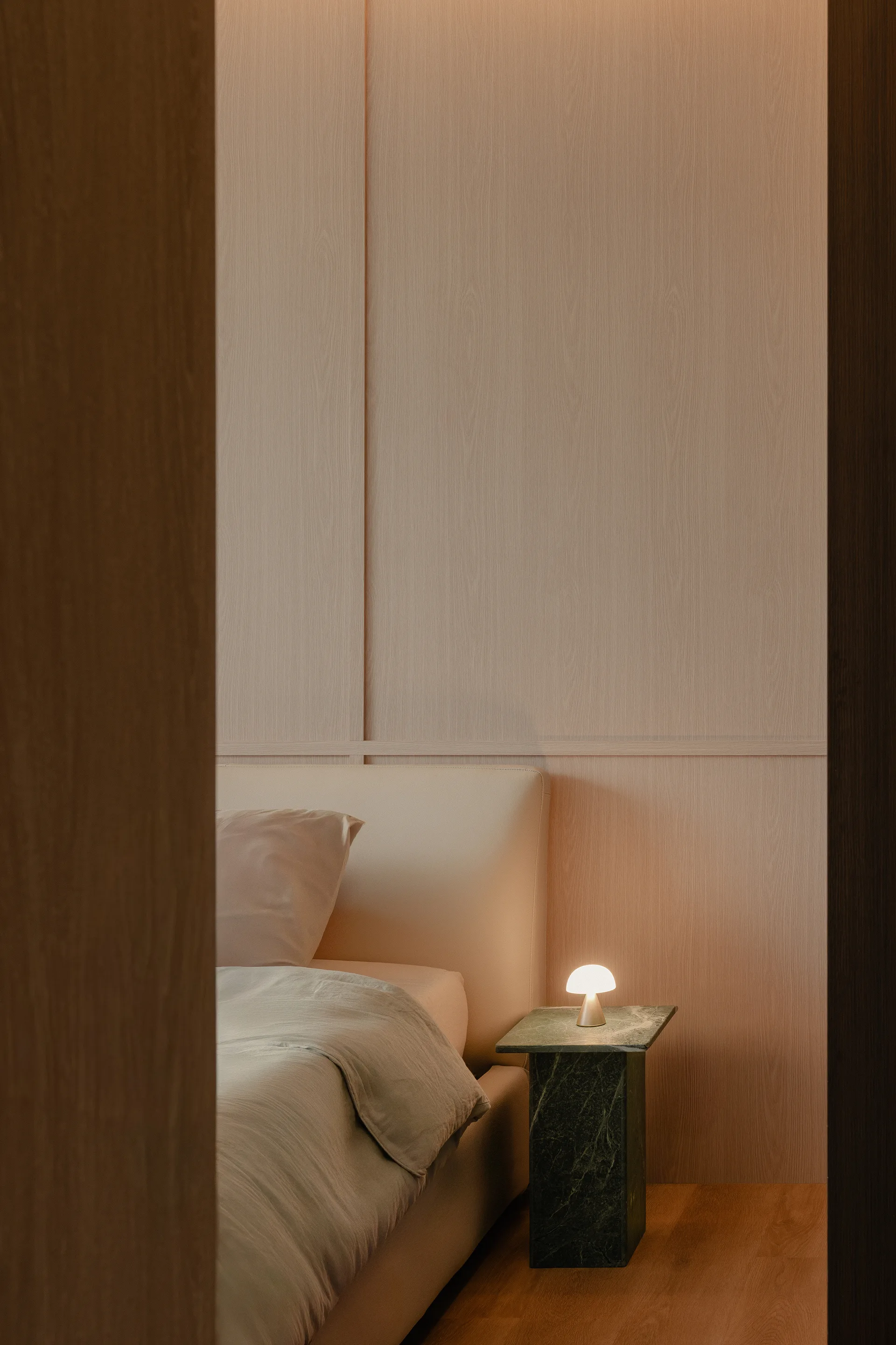 A minimalist bedroom featuring a beige upholstered bed and a glowing lamp on a dark green stone nightstand against wood paneling.