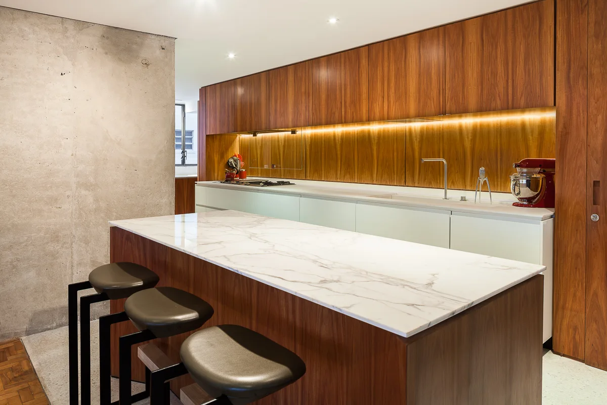 A modern kitchen featuring a marble-topped wood island with three bar stools, rich wood cabinetry, under-cabinet lighting, and an exposed concrete wall.