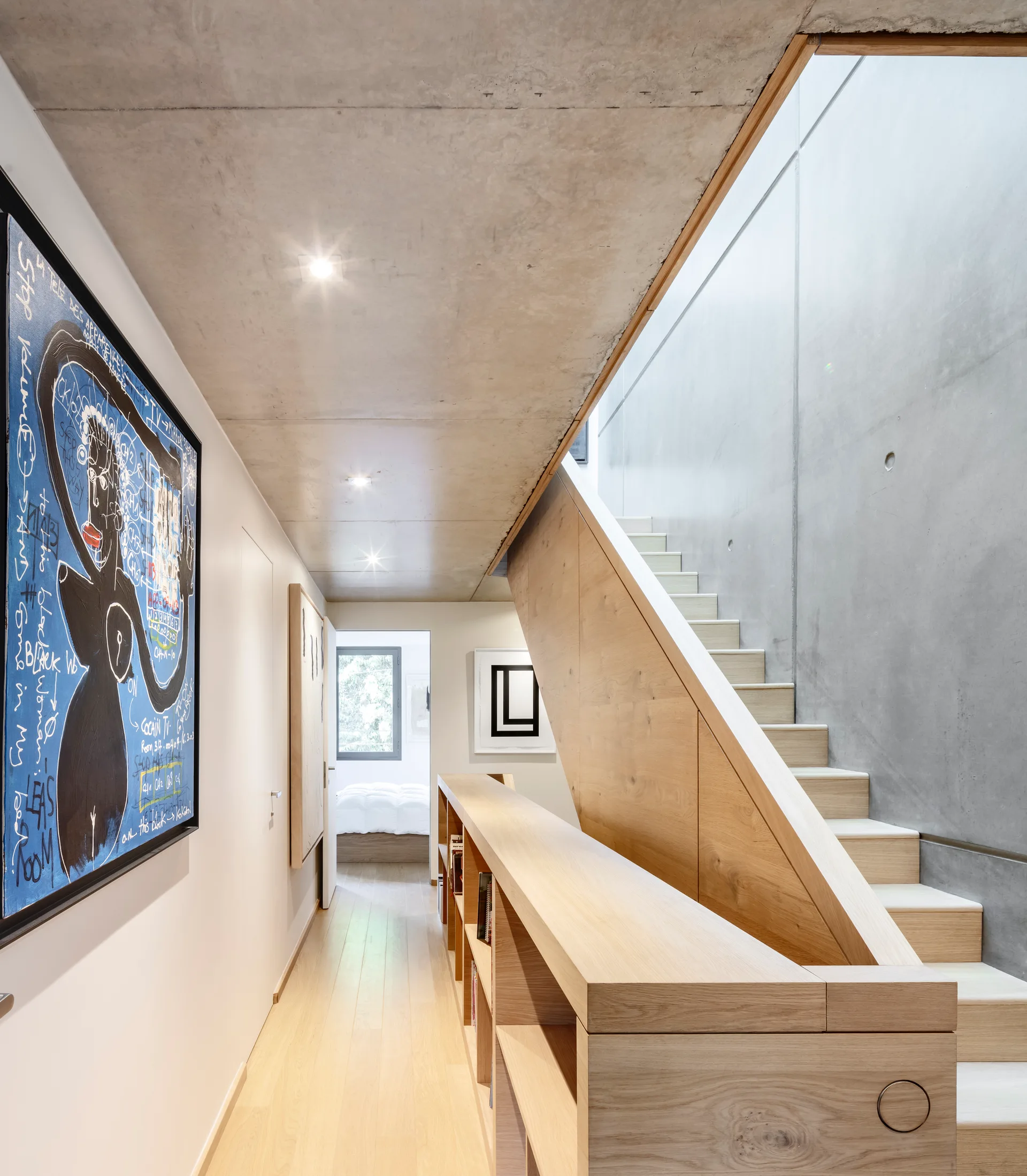 A minimalist hallway features an exposed concrete ceiling, a light wood staircase, a low wooden bookshelf, and framed artwork on the wall.