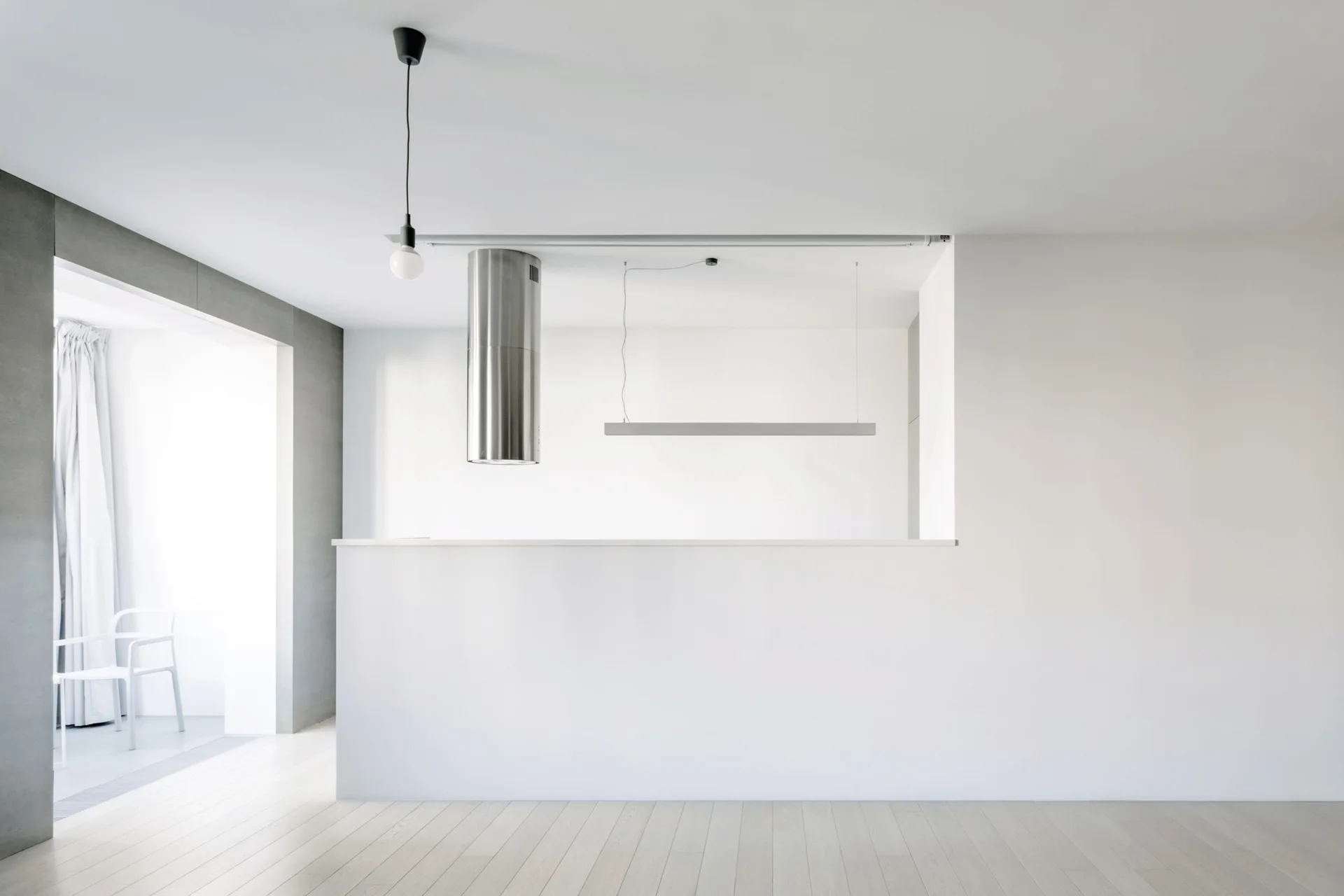 A minimalist kitchen features a solid white partition wall, light wood flooring, a stainless steel cylindrical range hood, and modern pendant lights.