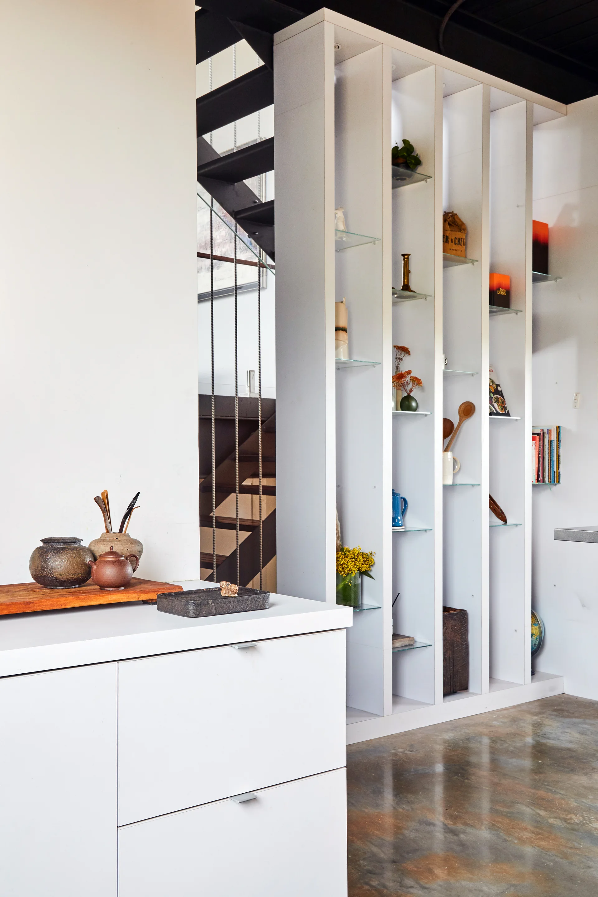 A tall white partition shelving unit with glass shelves stands next to an industrial black metal staircase and a polished concrete floor.