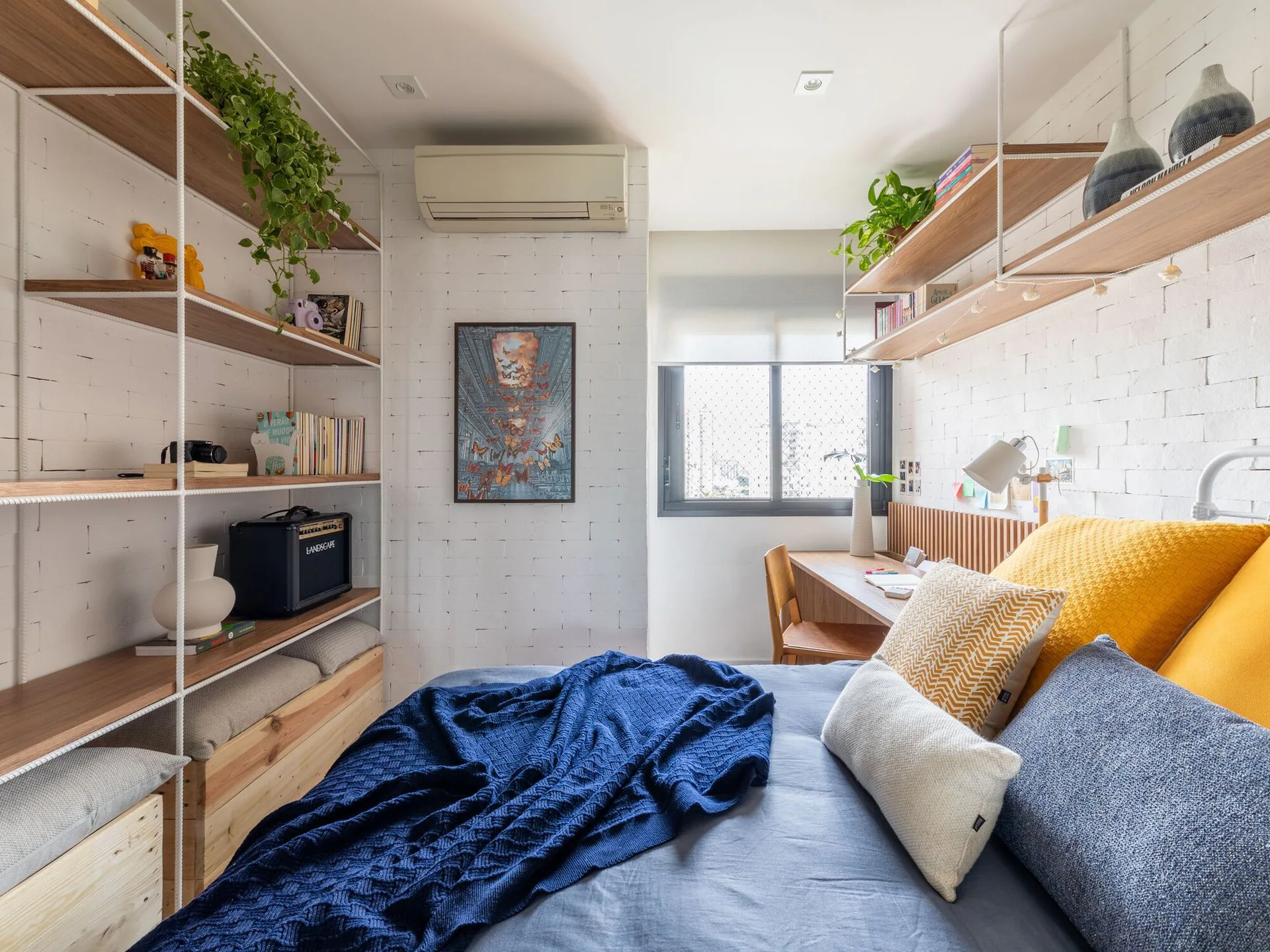 A small bedroom features a bed with blue bedding, white painted brick walls, open wooden shelves, and a desk under the window.