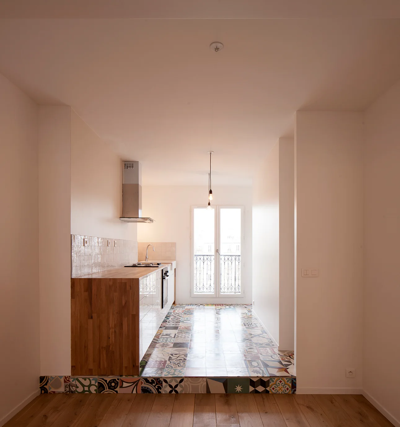 A galley kitchen on a raised platform features patterned encaustic floor tiles, a wood-topped counter, and a French balcony door.