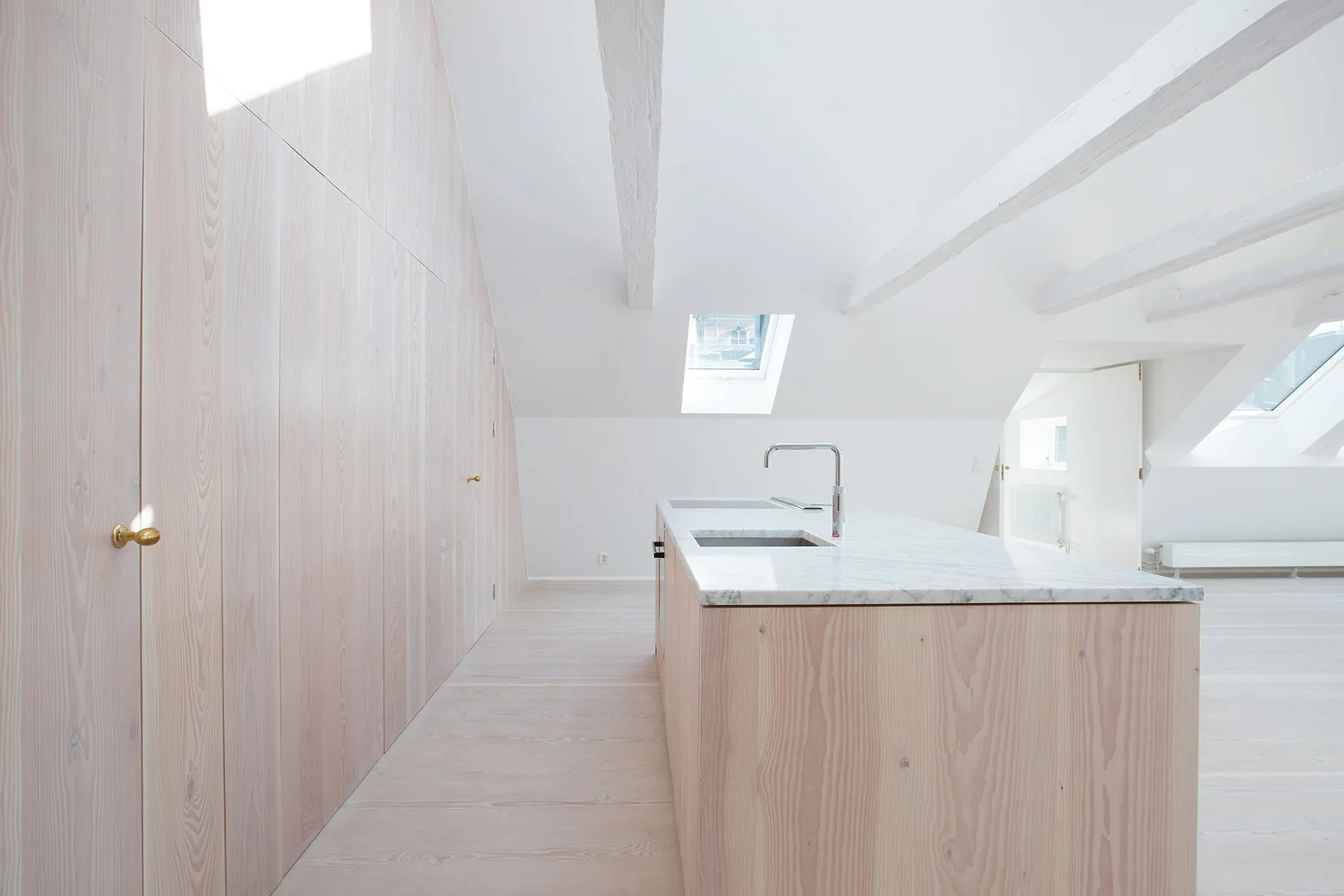 A minimalist attic kitchen featuring a light wood island with a marble countertop, integrated wood cabinetry, and vaulted white ceilings with beams.
