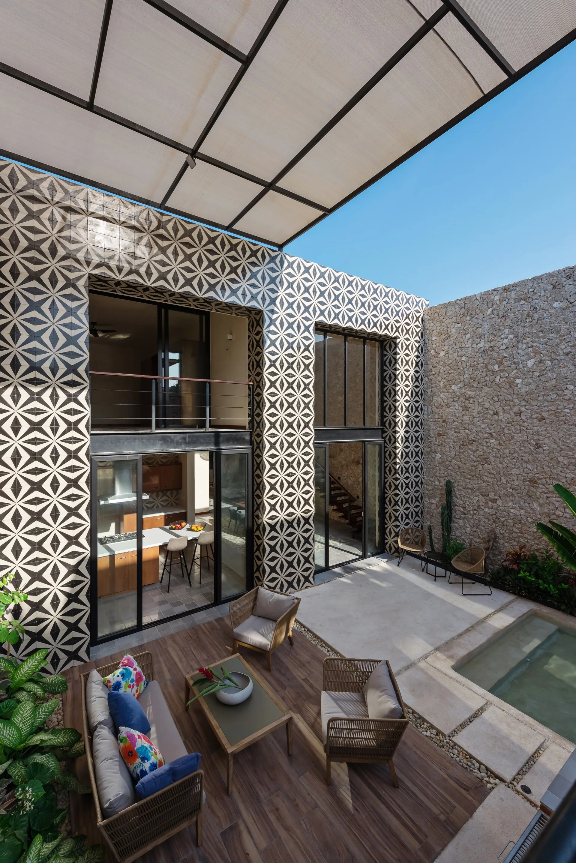A modern outdoor patio features wicker furniture, a rectangular plunge pool, a natural stone wall, and a black and white geometric tiled facade.