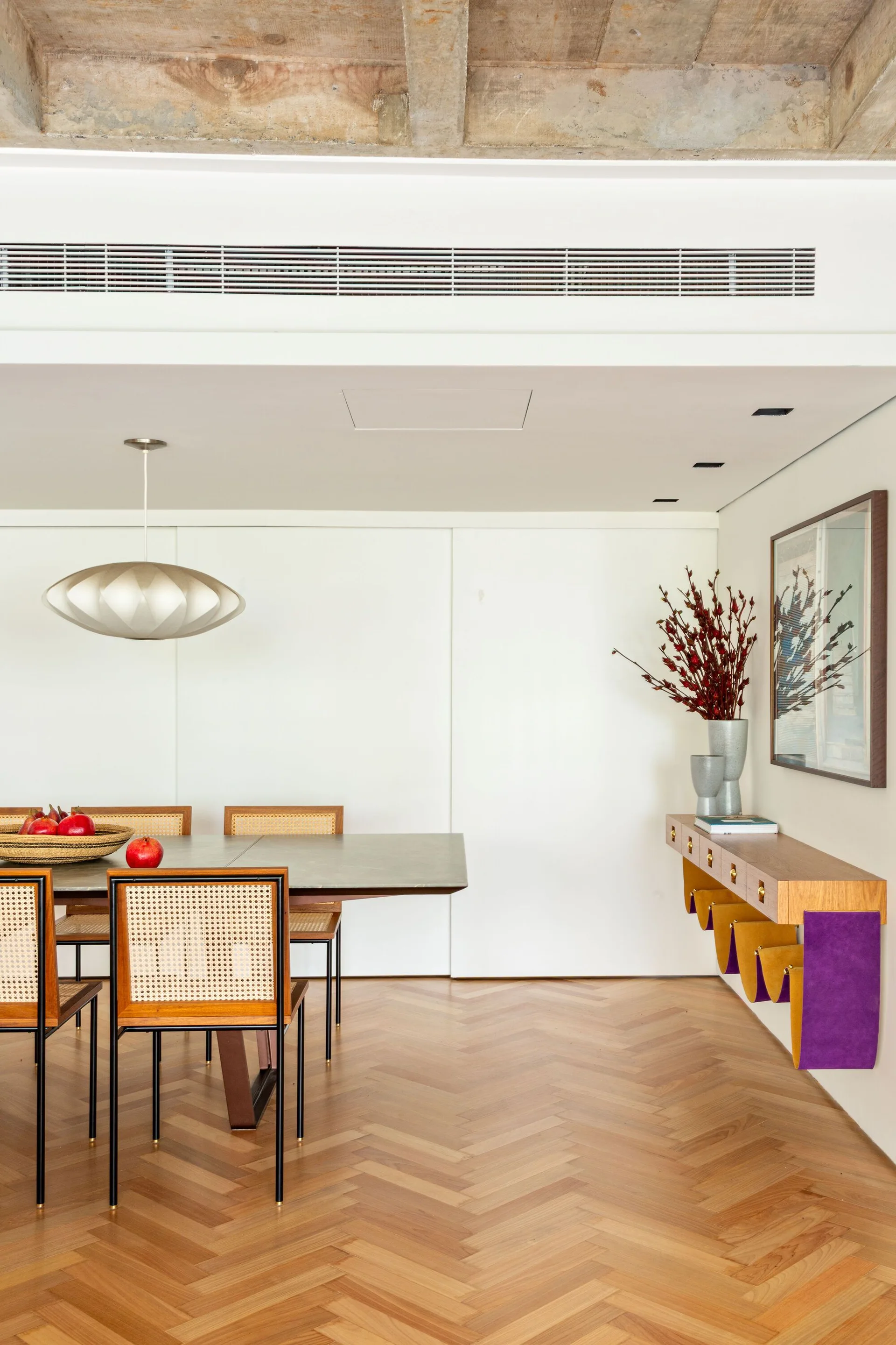 A dining room features a wood table with cane back chairs, a floating console, and a herringbone pattern wood floor beneath an exposed concrete ceiling.