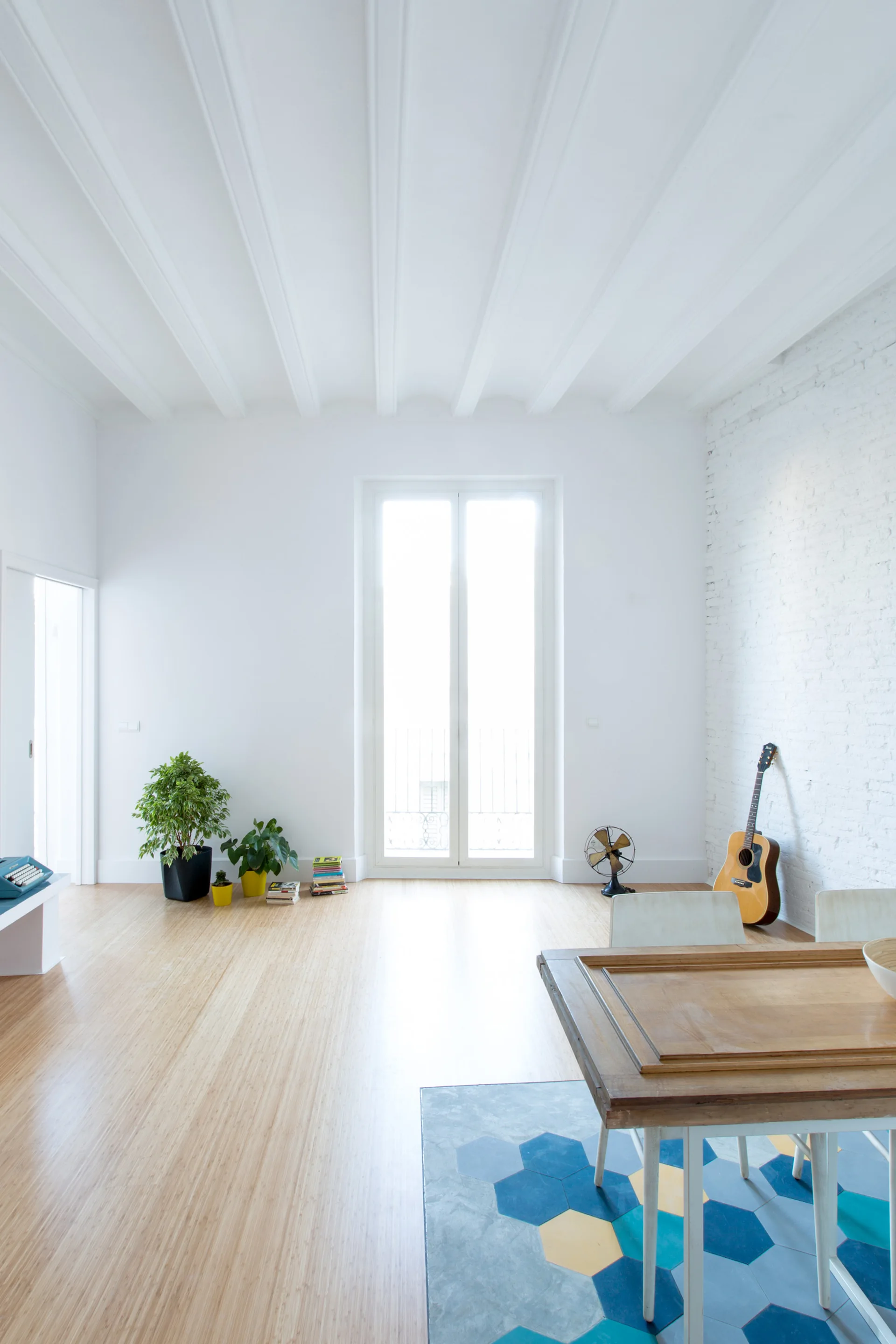 A bright dining space with white ceiling beams, a wooden table over colorful geometric floor tiles, and a white brick wall.