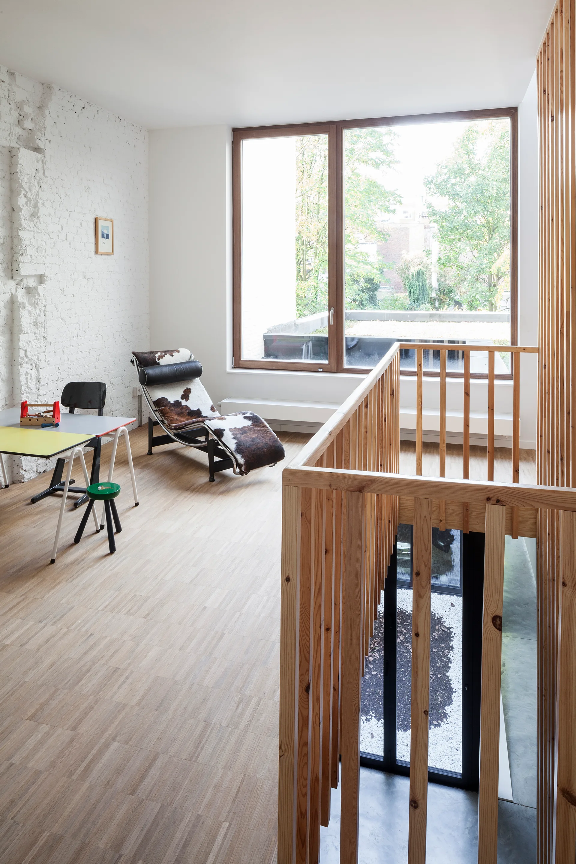 A light-filled upstairs landing featuring a wooden slatted staircase railing, a cowhide chaise lounge, and a large window.