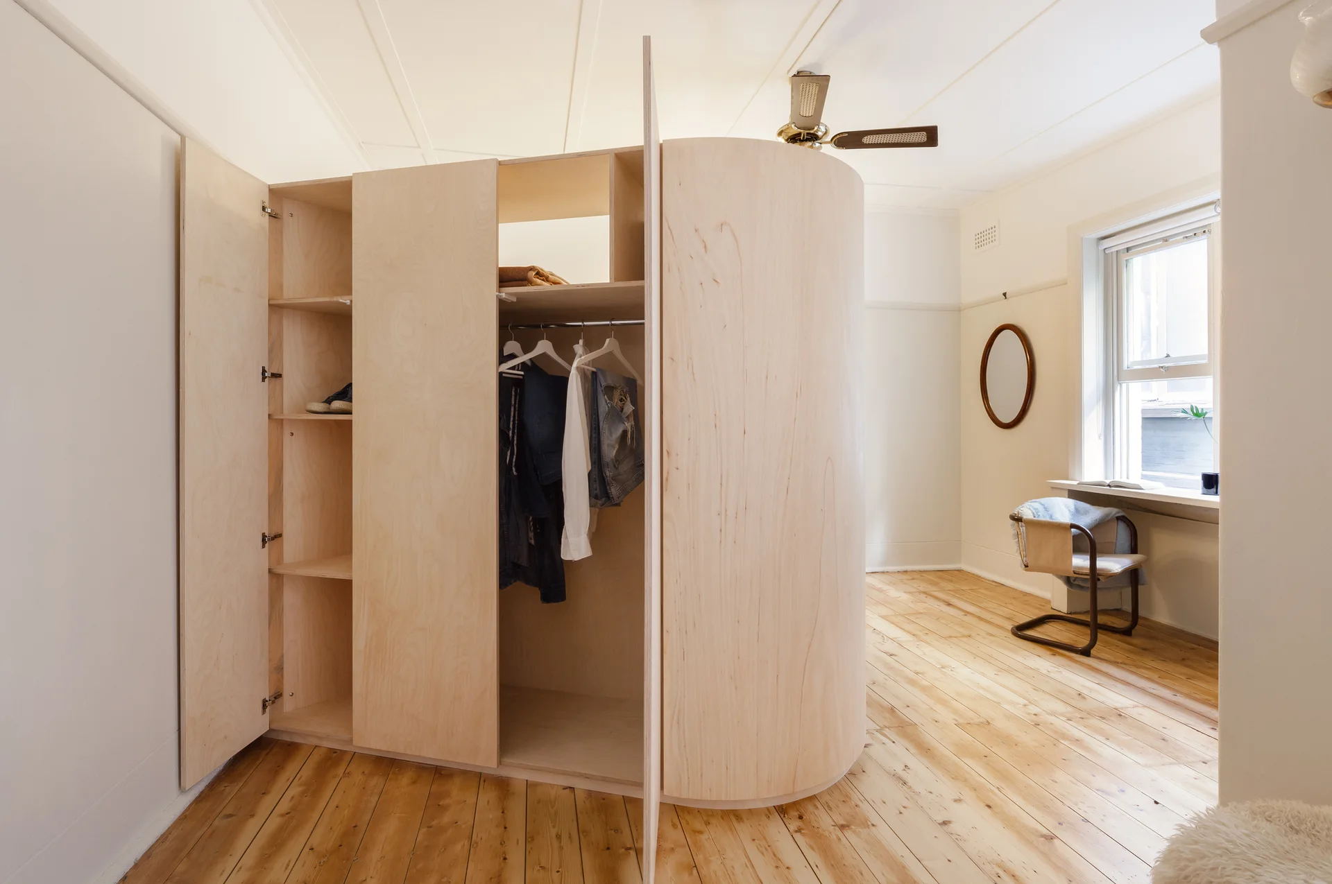 A light wood curved plywood wardrobe stands open in a minimalist bedroom with pine floorboards and a desk by the window.