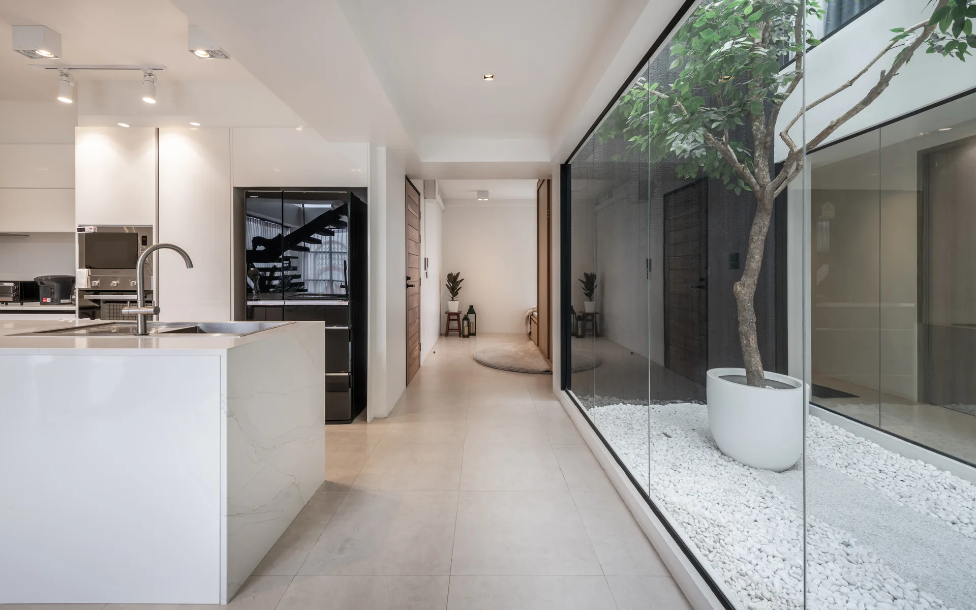 A minimalist white kitchen island and black refrigerator stand next to a hallway lined by a glass-walled courtyard with white gravel and a potted tree.