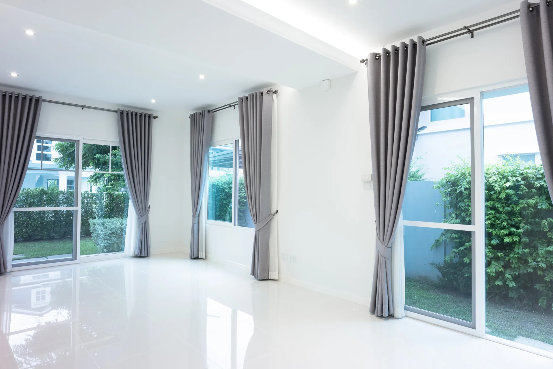 An empty room with glossy white tile flooring, white walls, and multiple sliding glass doors decorated with long grey drapes.