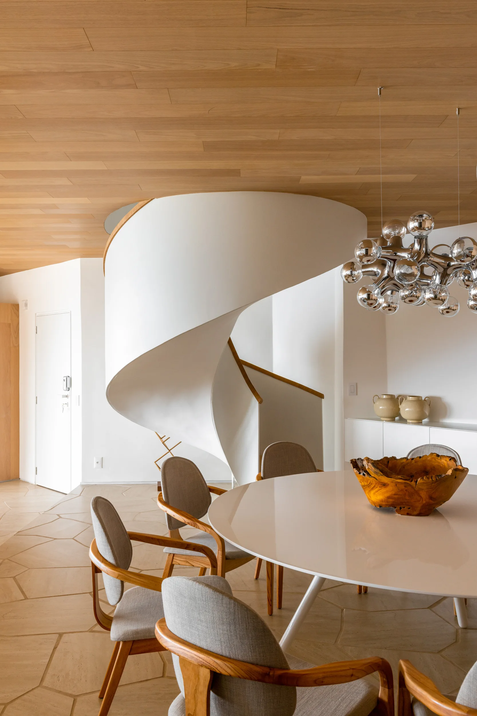 A modern dining room features a round white table, wood-frame chairs, a white spiral staircase, and a wood plank ceiling.