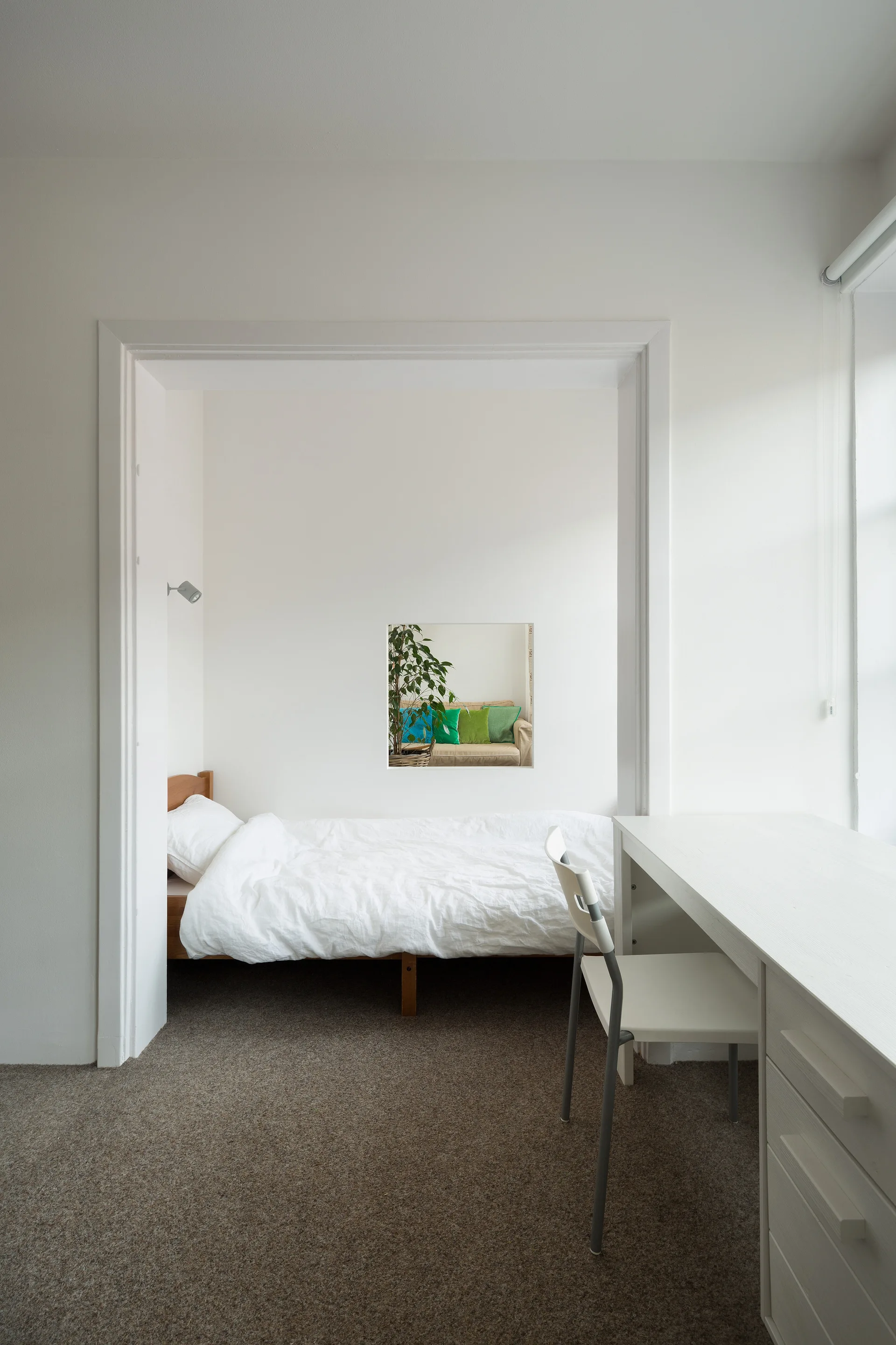 A minimalist bedroom features a bed tucked into an alcove next to a white desk, with a square wall cutout opening to an adjacent room.