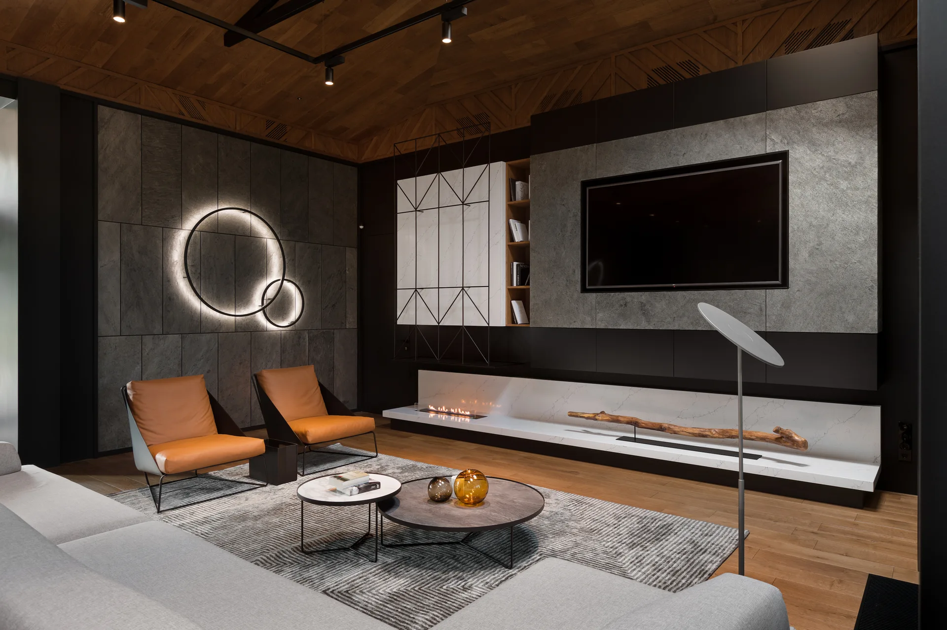 A moody modern living room features a grey sectional sofa, tan leather chairs, a backlit circular wall light, and a stone-clad television wall.