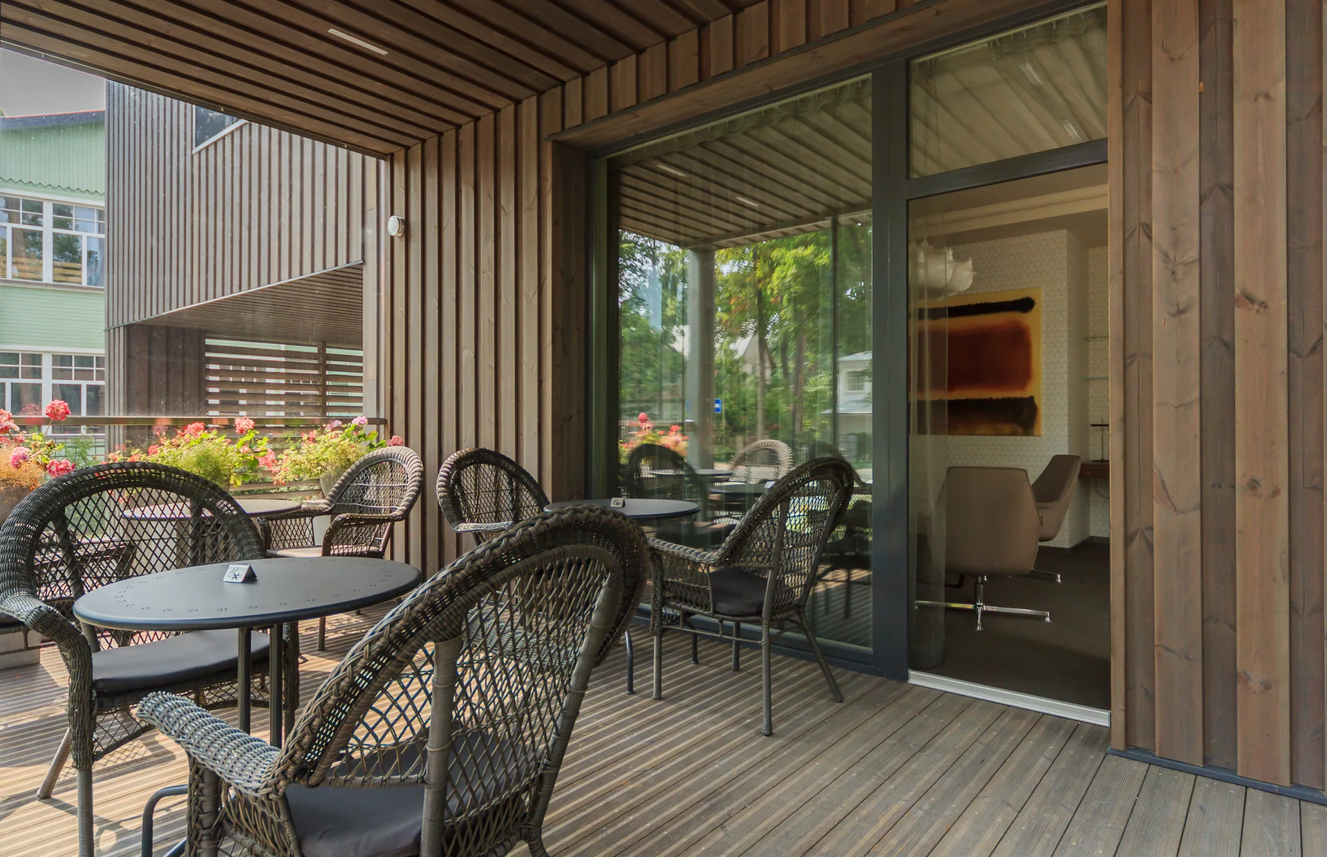 Dark wicker chairs and tables sit on a wooden plank deck under a slatted timber ceiling with large glass doors leading inside.