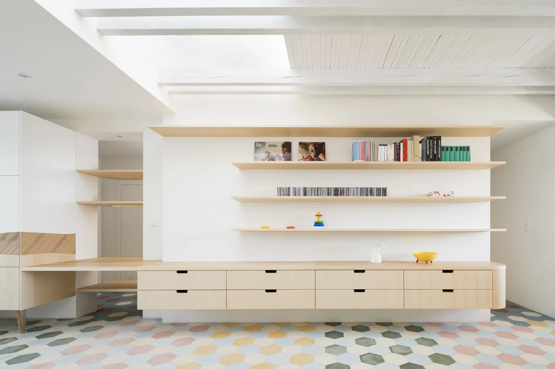 A minimalist room features a long light wood built-in drawer unit, floating shelves, and a colorful hexagonal tiled floor under a white beam ceiling.
