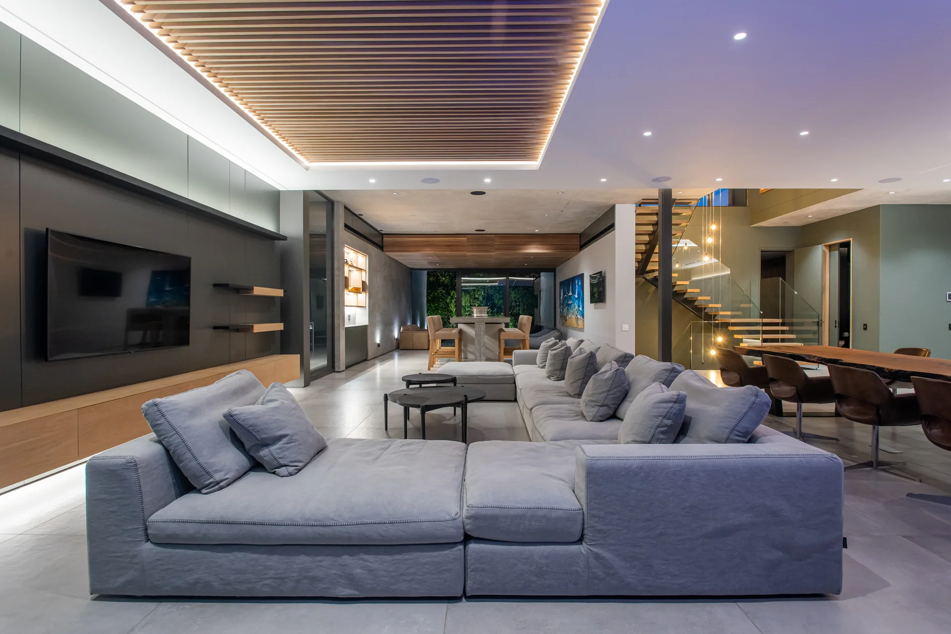 A spacious modern living room features a large grey sectional sofa, a decorative wood slat ceiling, and a floating staircase with glass railings.