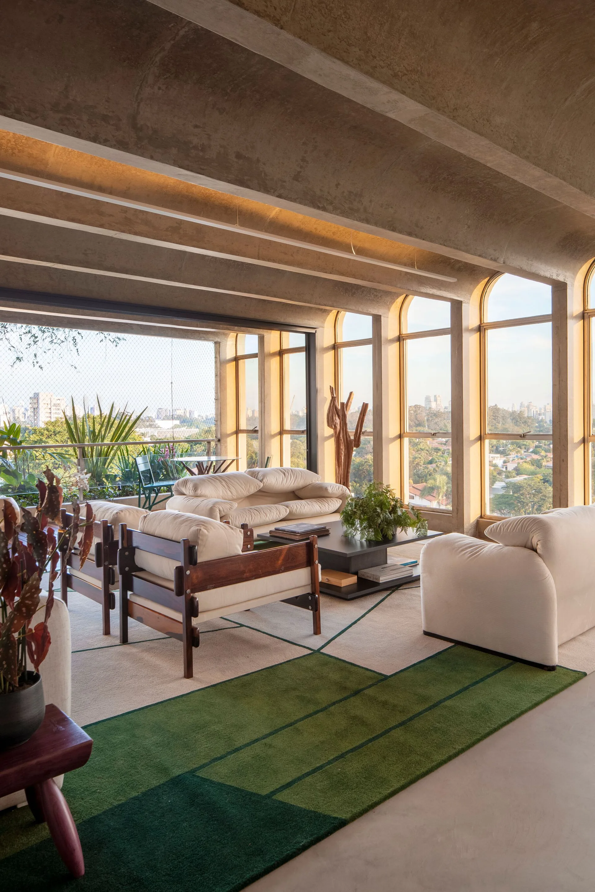 A sunlit living room features plush off-white sofas, a low coffee table, and a green-blocked rug beneath a vaulted concrete ceiling and tall arched windows.