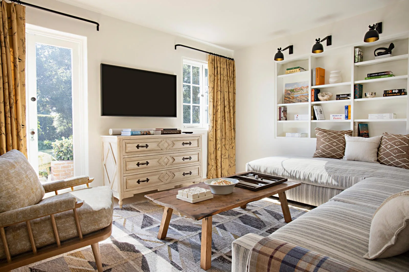 A living room features a striped sectional sofa, a low wood coffee table, patterned curtains, and a recessed white shelving unit filled with books.