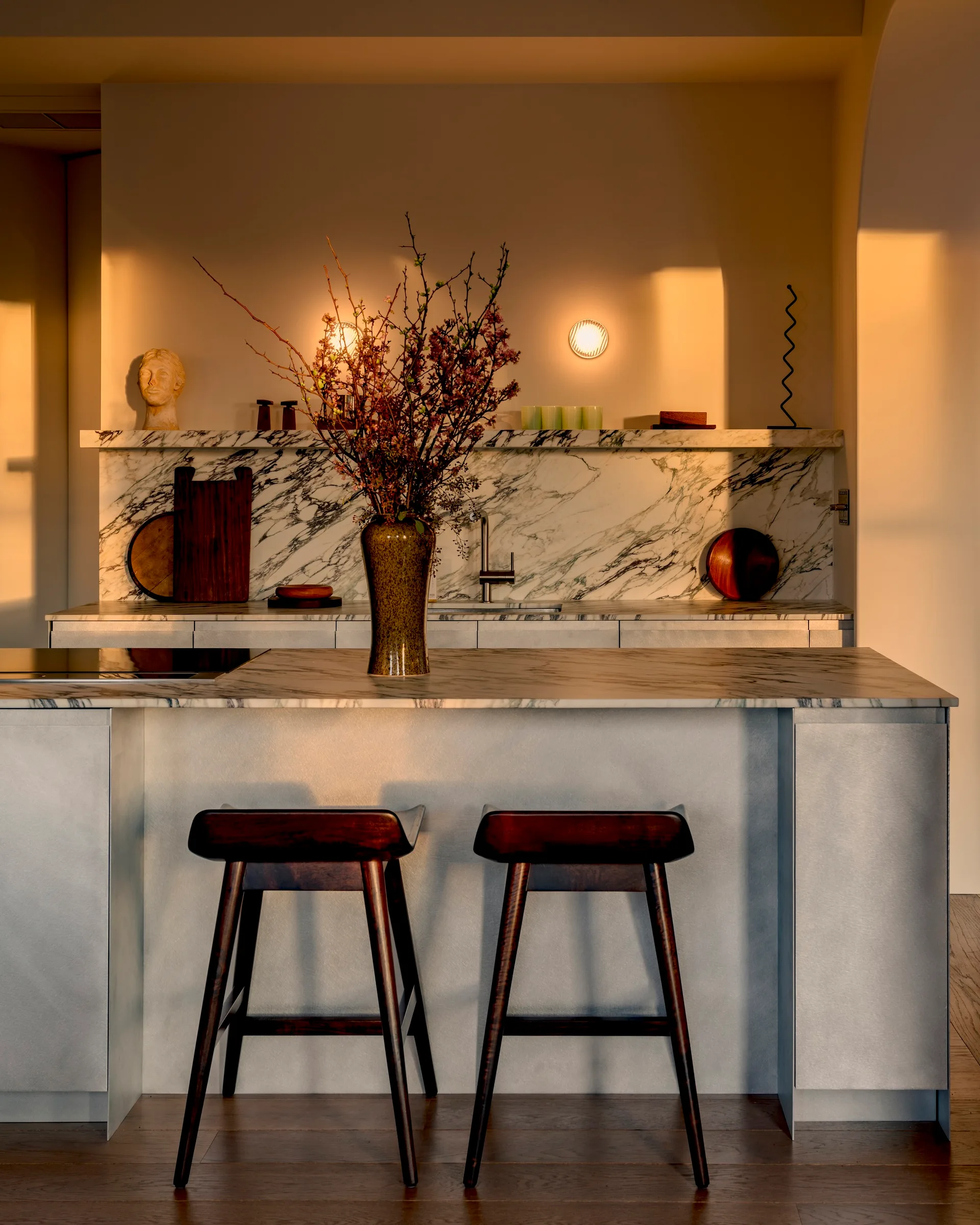 A minimalist kitchen features a veined marble backsplash and island, accented by two dark wood bar stools and glowing wall sconces.