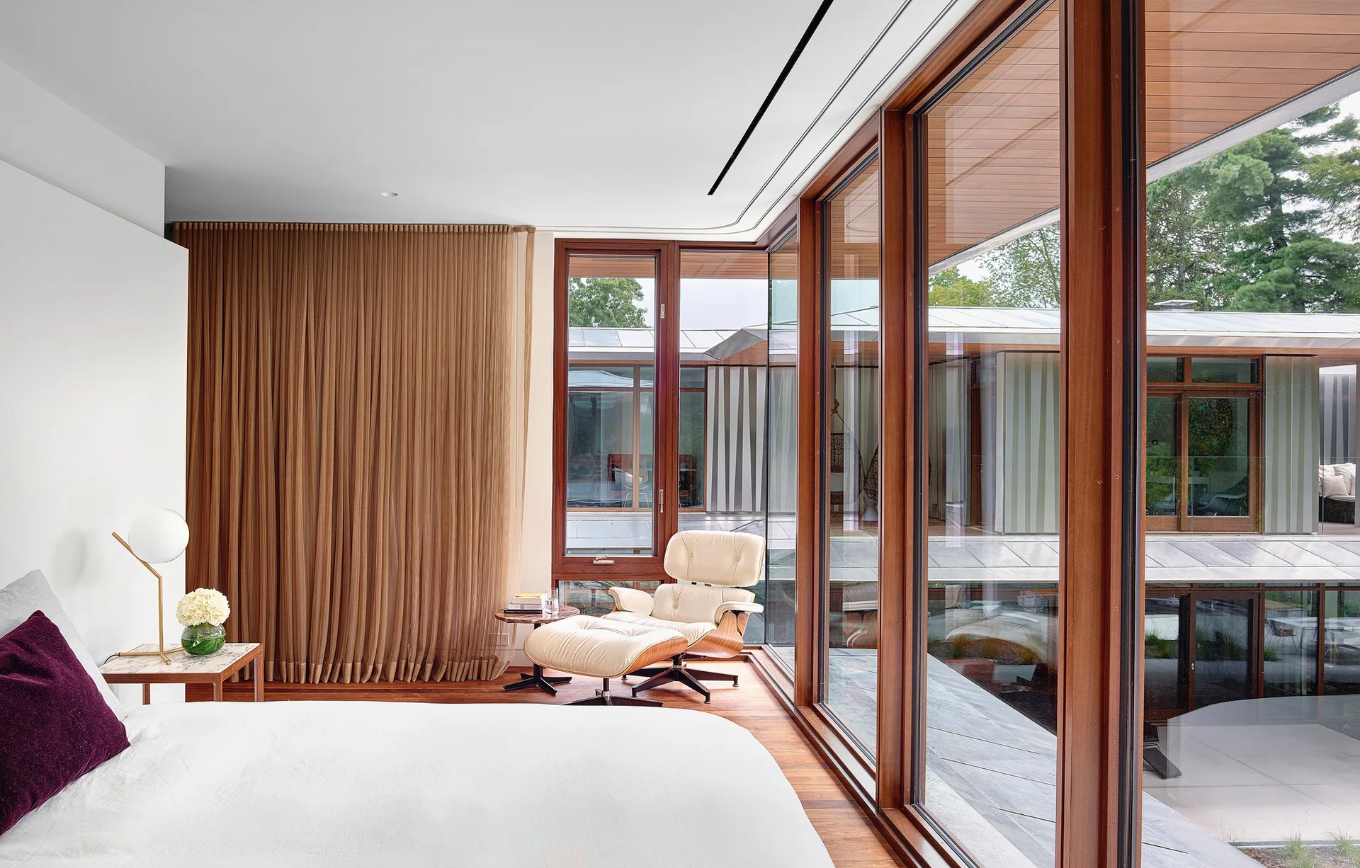 A modern bedroom with a cream leather lounge chair and ottoman, large wood-framed glass windows, and sheer brown curtains.