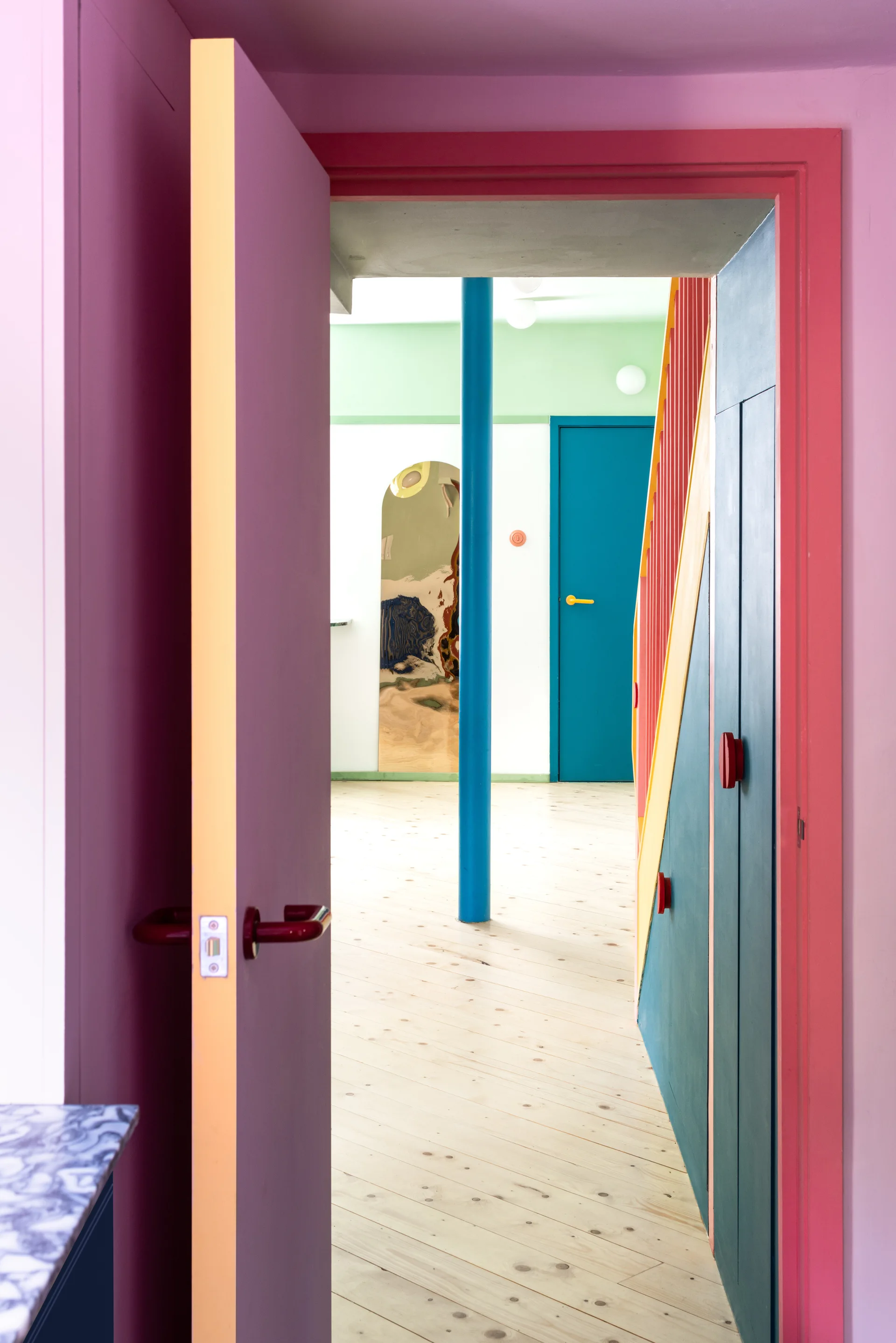 An open pink door looks into a hallway with a blue column, light wood flooring, green ceiling, and teal staircase paneling.