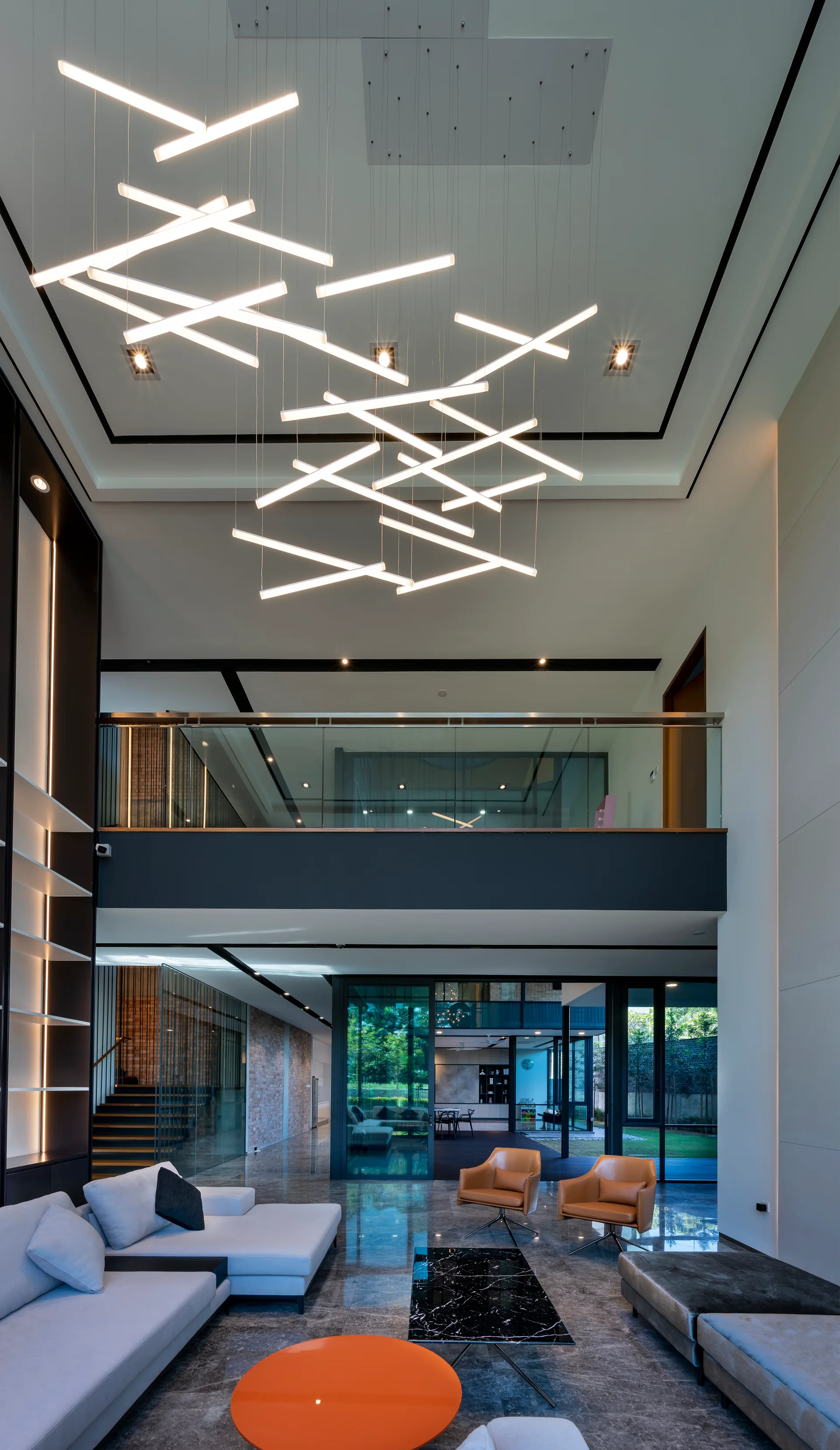 A double-height living room features a large intersecting linear chandelier, a grey modular sofa, tan leather armchairs, and reflective grey tile floors.