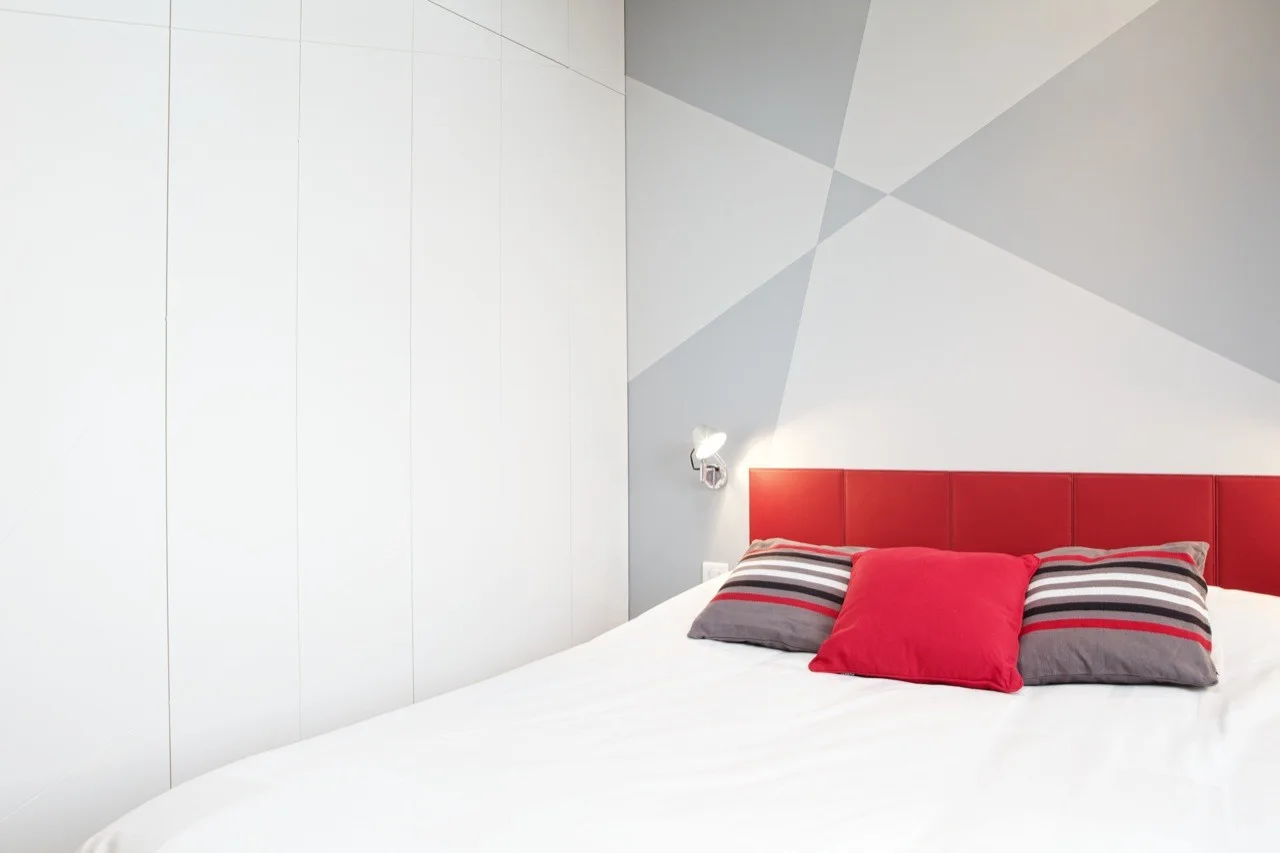 A modern bedroom features a red upholstered headboard, striped pillows, a gray geometric accent wall, and floor-to-ceiling white closets.